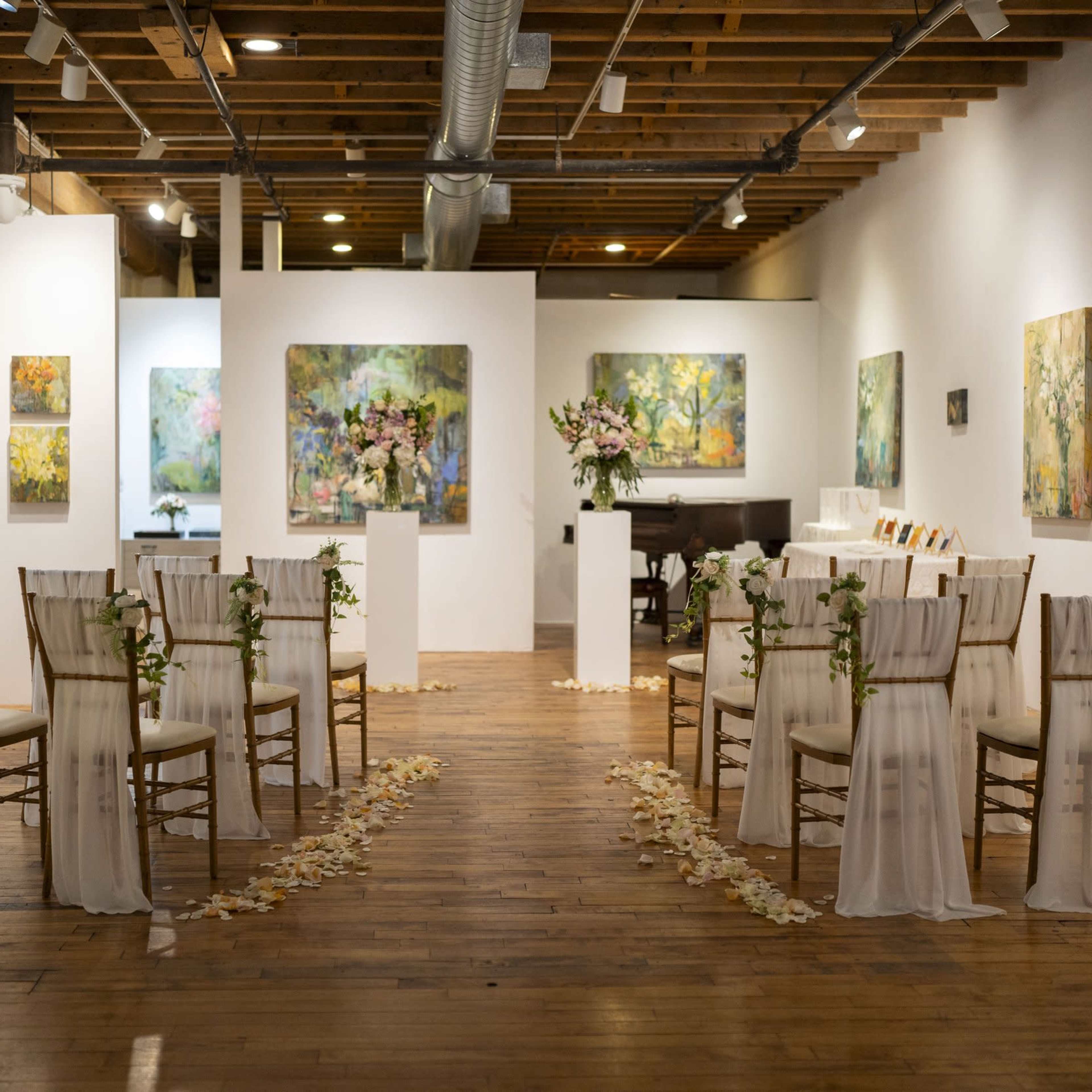 A wedding ceremony set up in an art gallery features rows of chairs adorned with white fabric and floral decorations, leading to a floral arrangement on a pedestal.