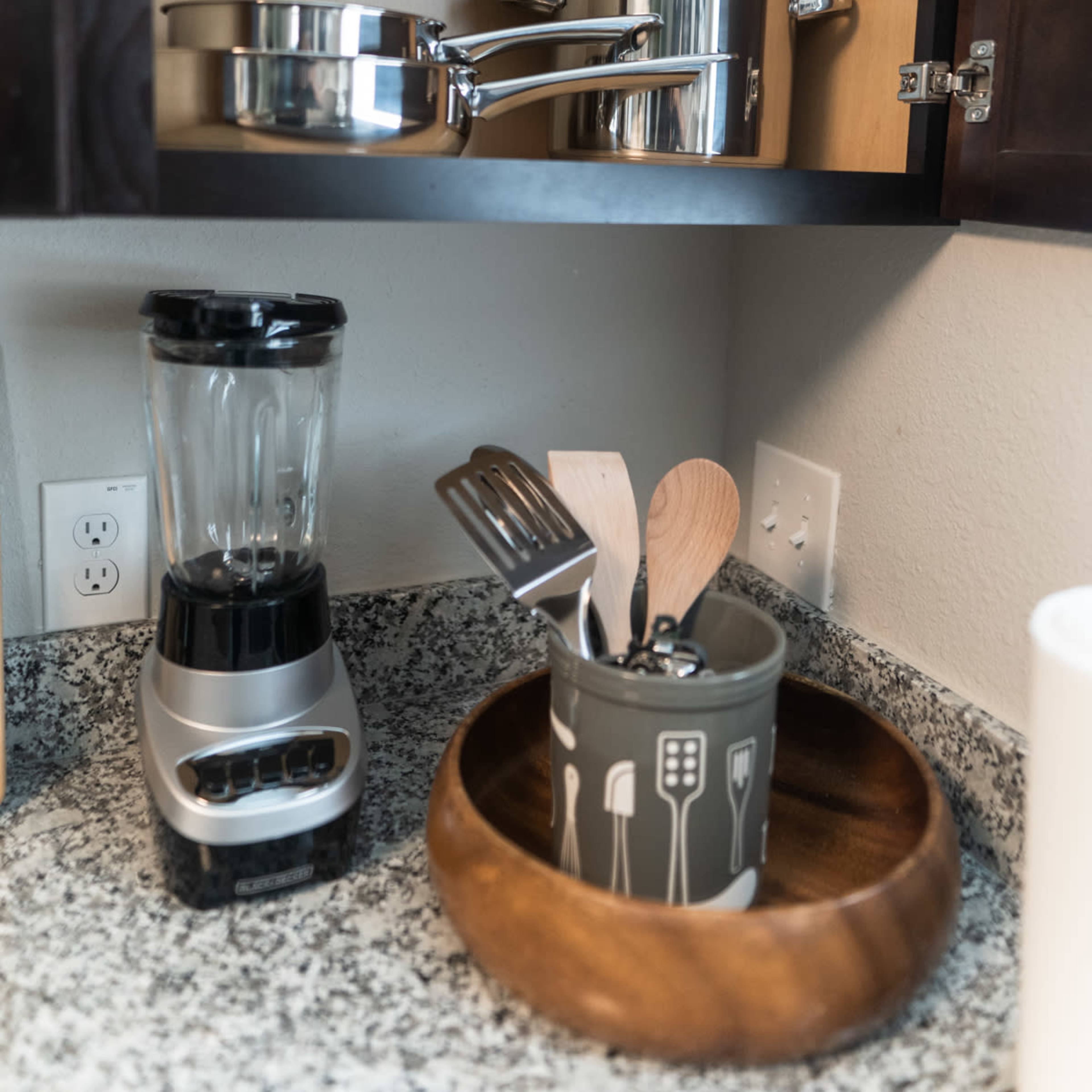 A kitchen cabinet displays metal cookware and a blender on a countertop next to a bowl containing wooden utensils.