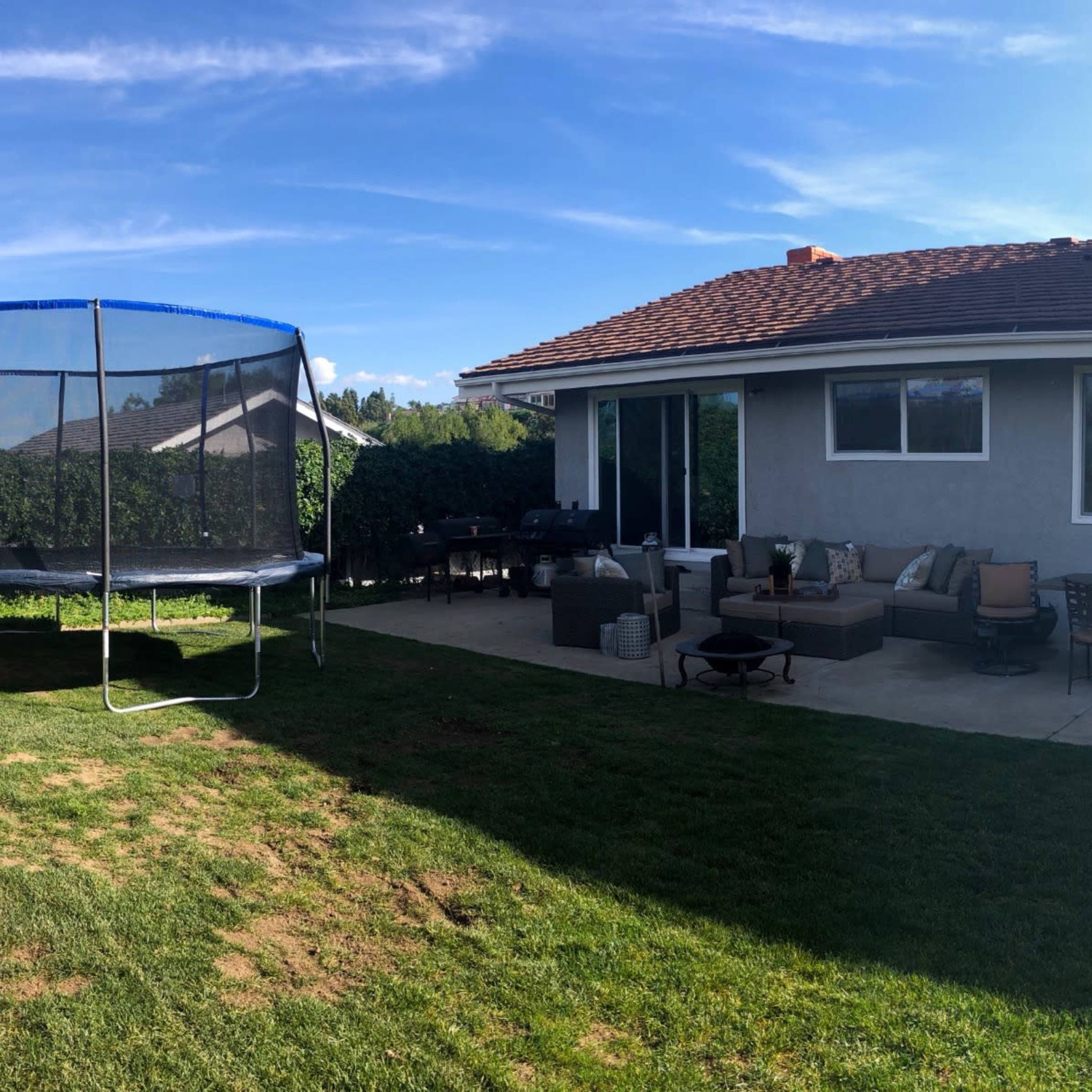 A suburban backyard features a paved patio with outdoor furniture, a trampoline, and a soccer goal, surrounded by grass and trees.