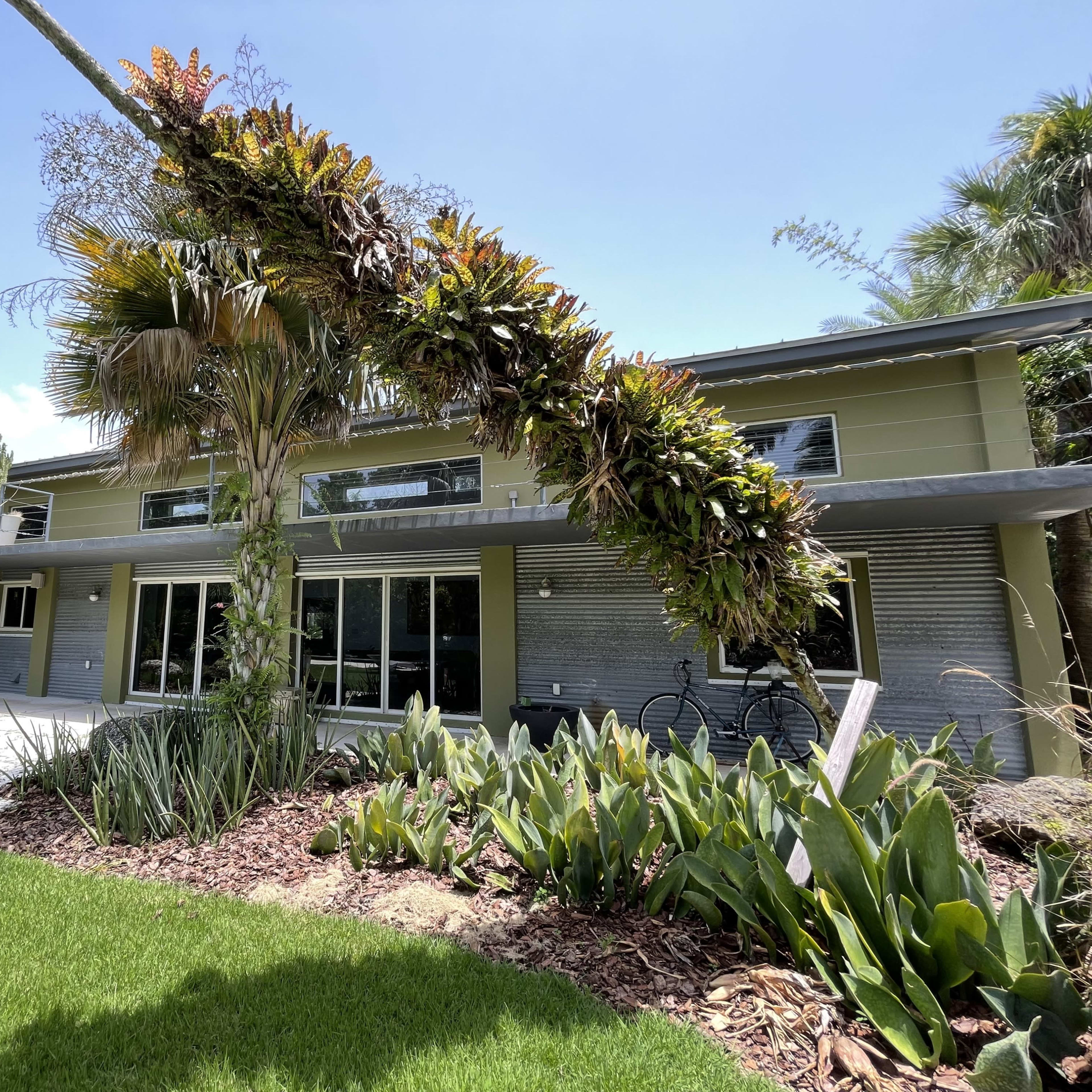 A modern house with large windows surrounded by tropical plants and landscaping.