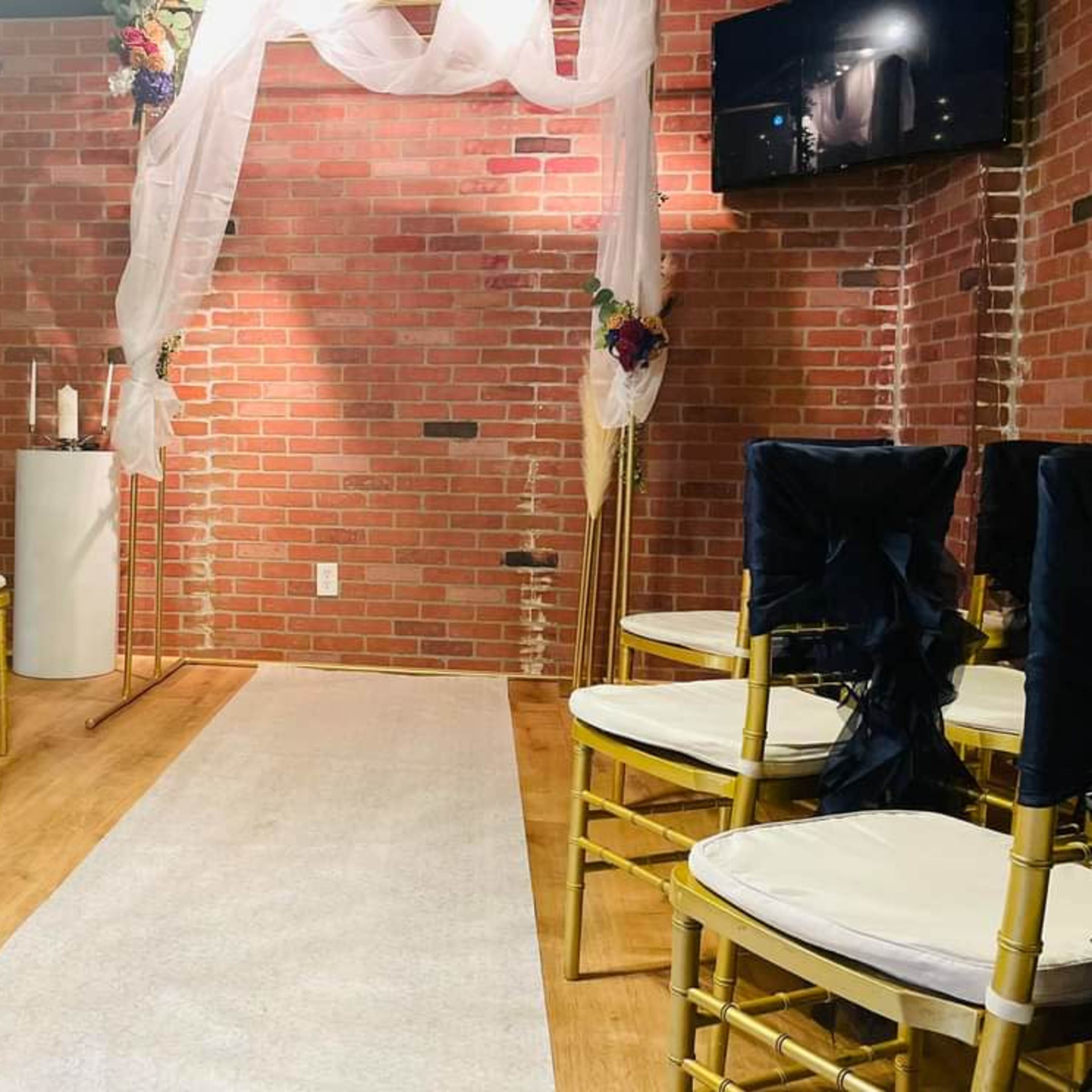 A small indoor ceremony space features rows of gold chairs with navy blue sashes facing a white aisle, adorned with fabric drapery and a small altar against a brick wall.