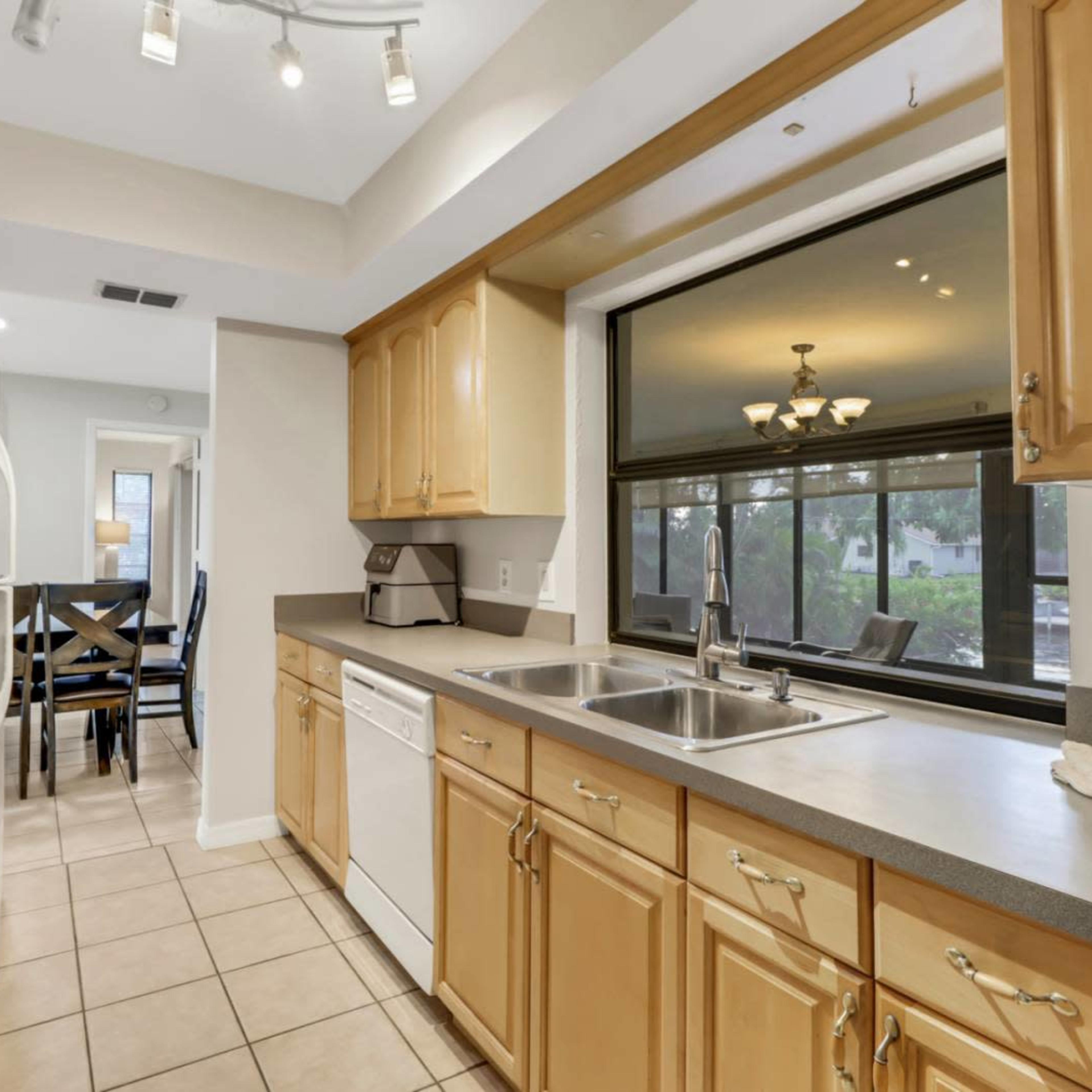 A clean kitchen features light wood cabinets, a double sink, and a view of a dining area through a large window.