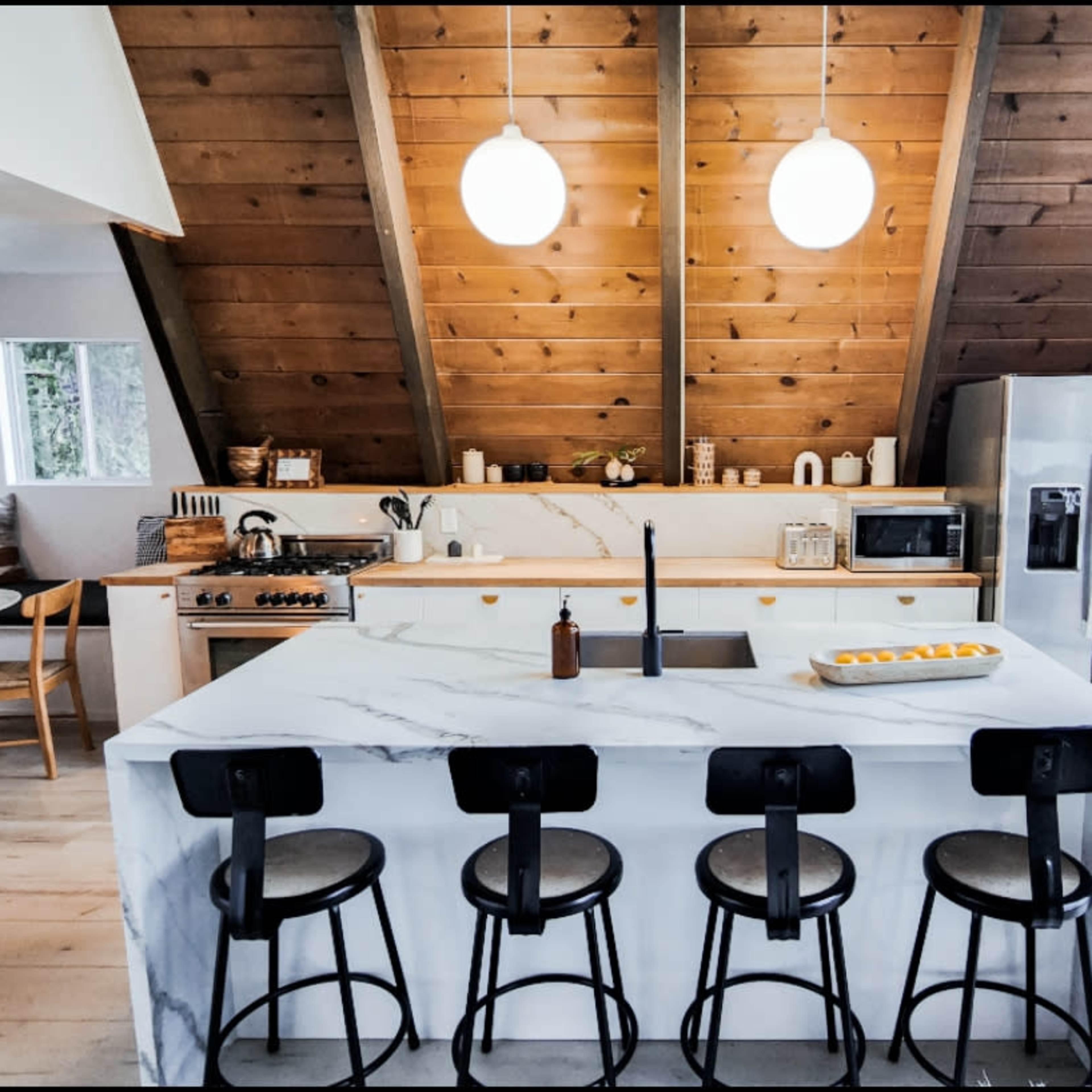 A modern kitchen with a marble island, four black stools, and a wooden sloped ceiling.