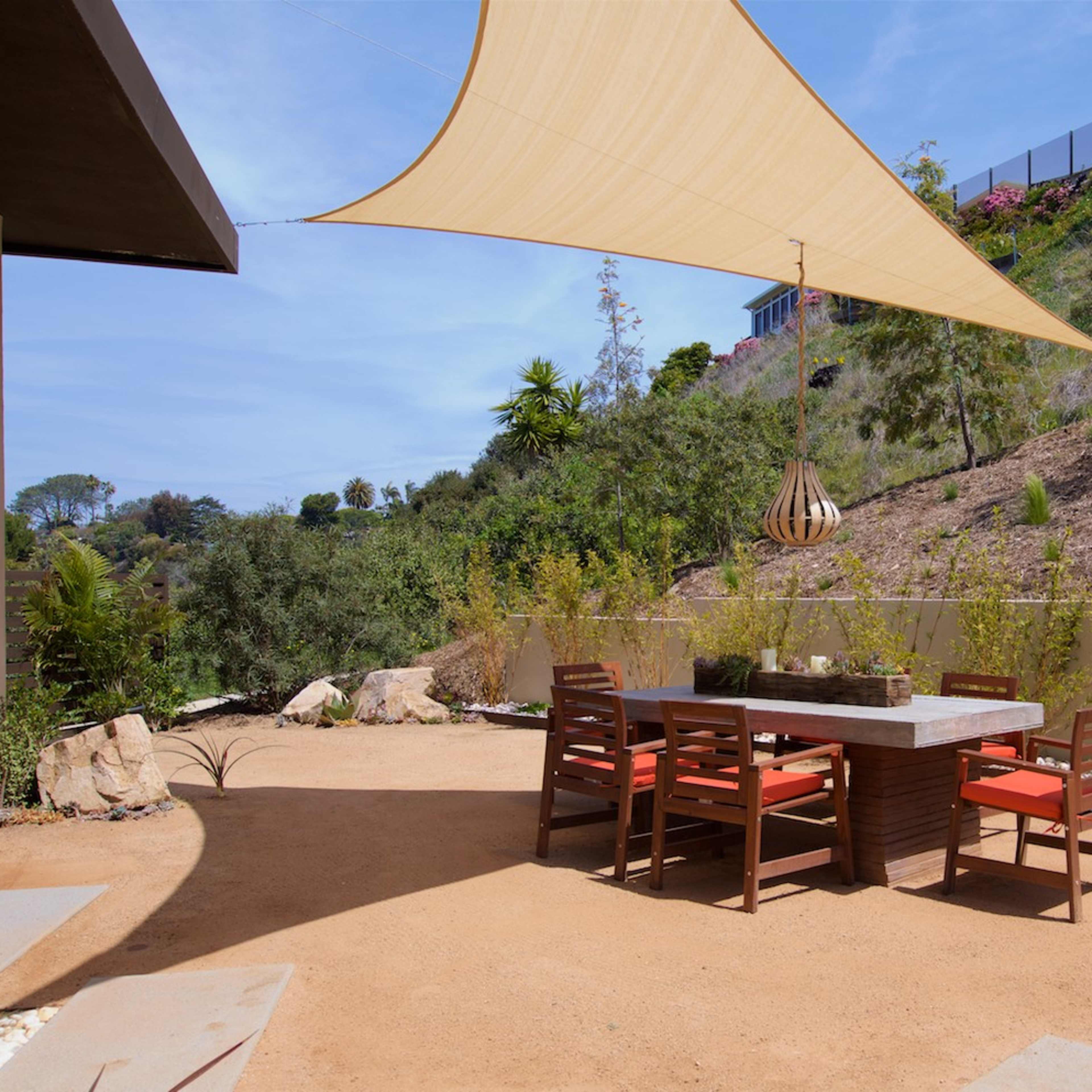 A shaded outdoor dining area features a stone table surrounded by wooden chairs on a sandy patio.