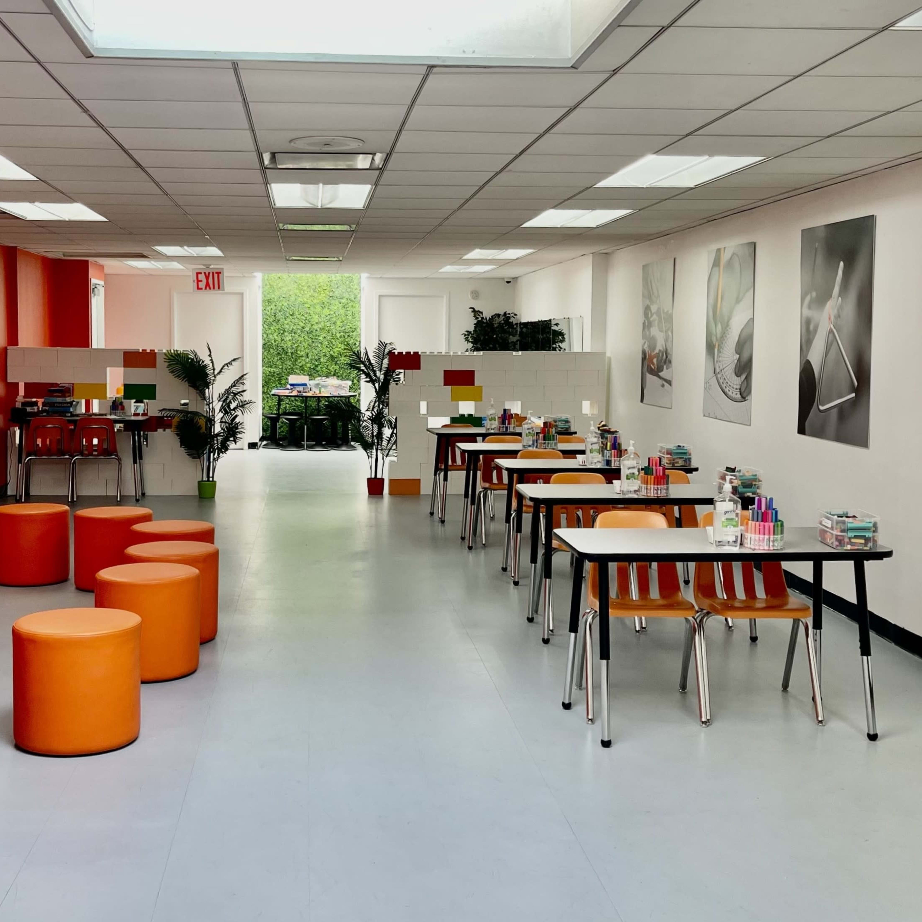 A modern workspace featuring orange-accented seating, several tables with supplies, and large windows providing natural light.