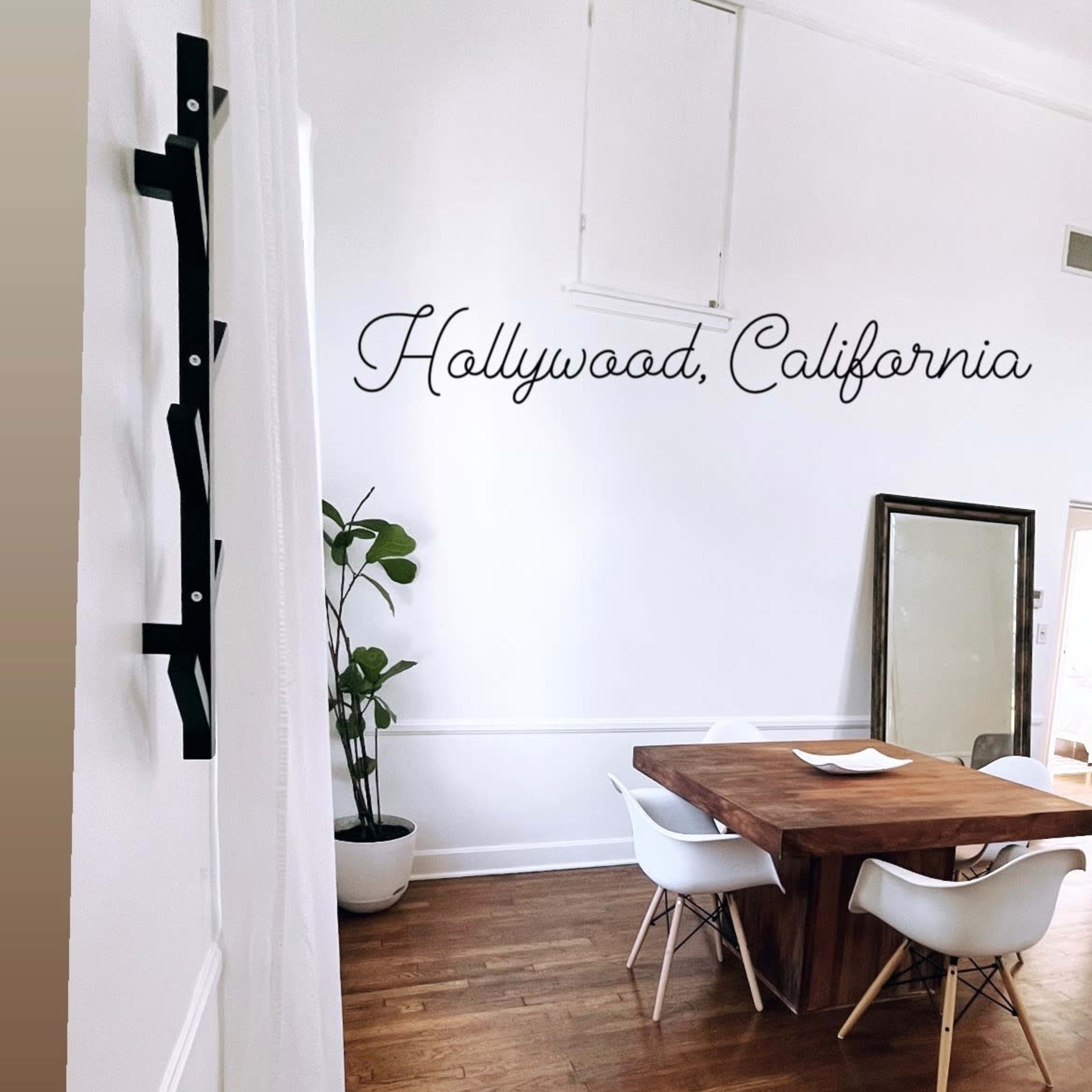 A dining area with a wooden table, white chairs, a large mirror, and a plant against a white wall featuring the text "Hollywood, California."