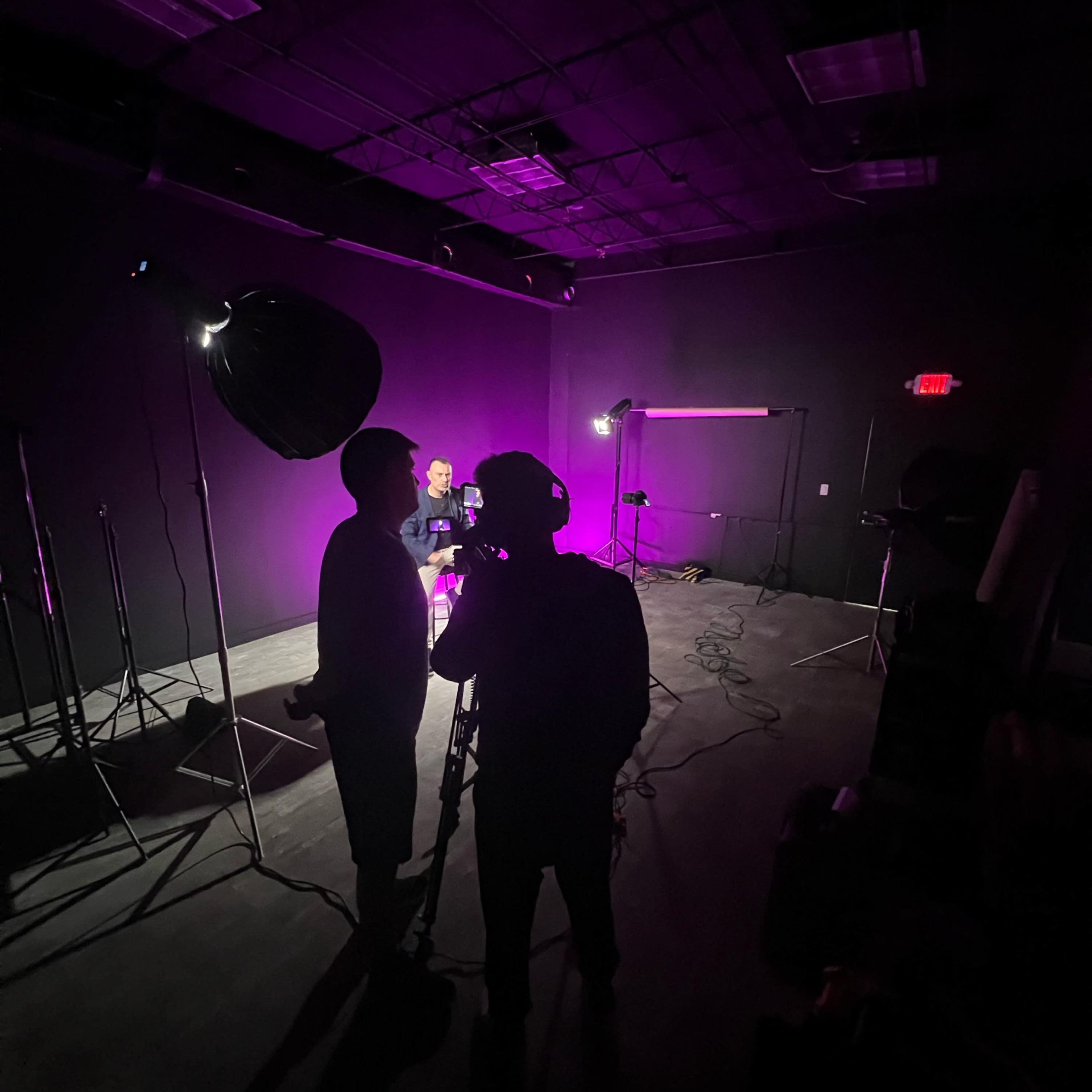 A dimly lit studio setup features two individuals engaged in conversation, with cameras and lighting equipment arranged nearby against a purple backdrop.