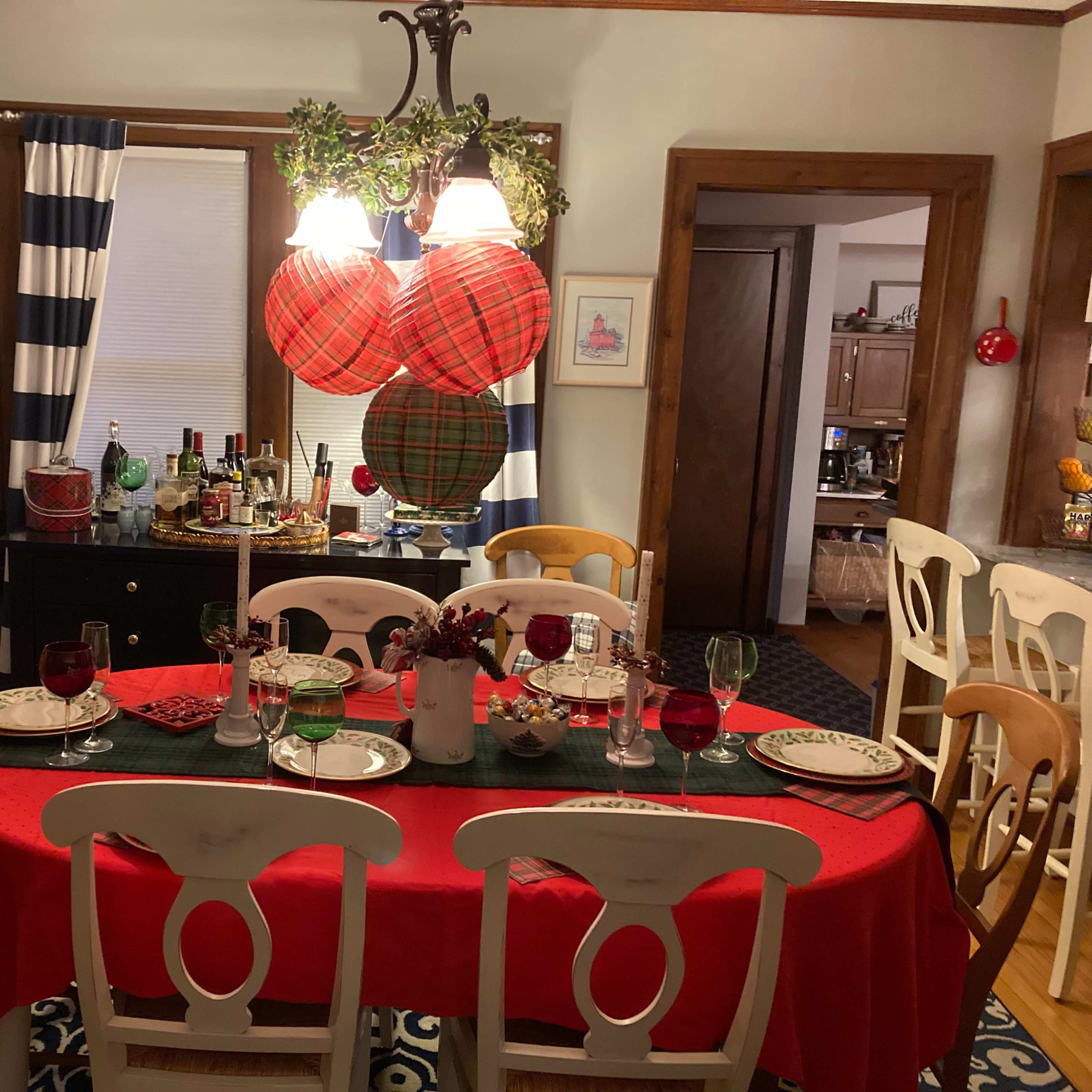 A dining room features a table set with red and green decorations, surrounded by white chairs, and adorned with a chandelier and checkered wall hangings.