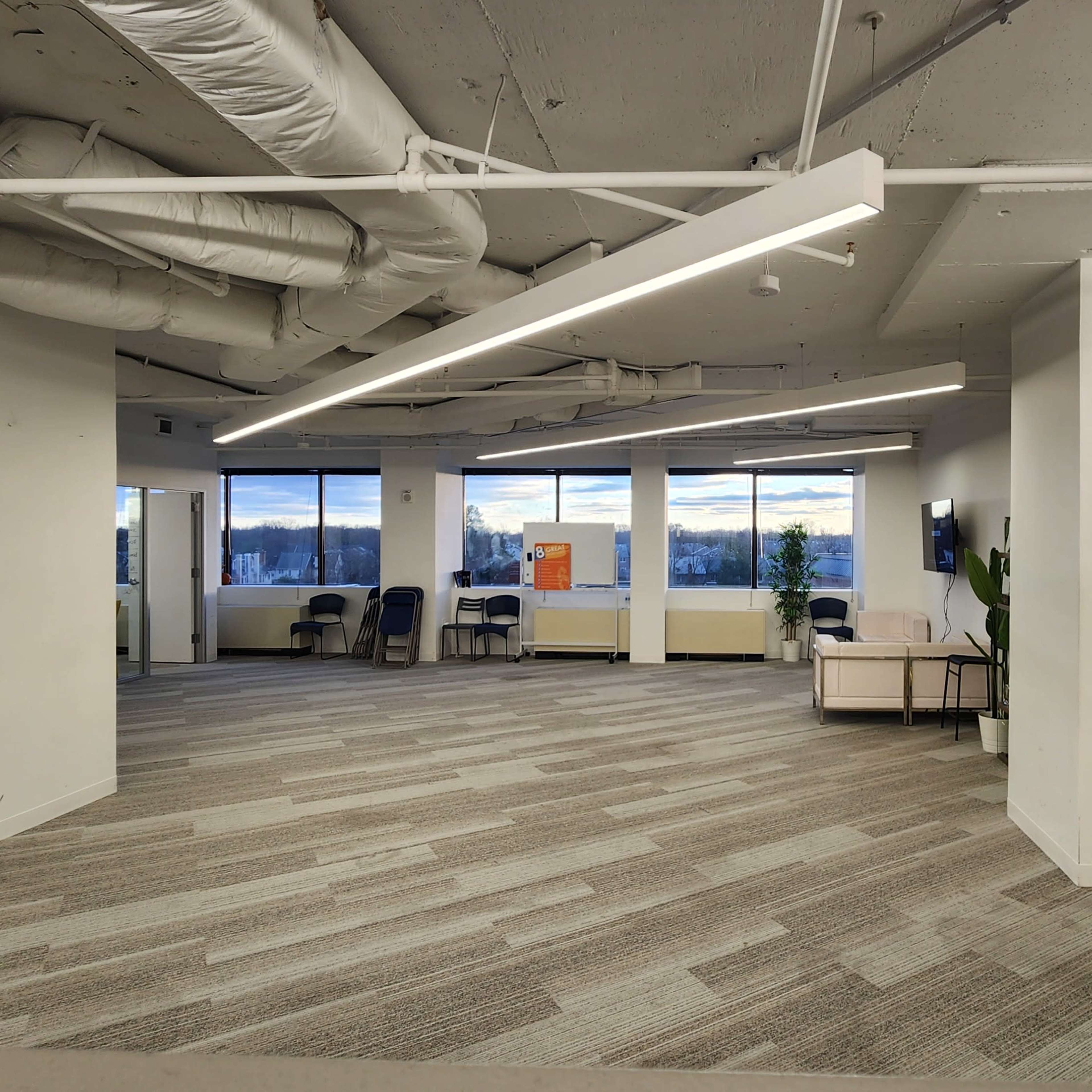 An office interior featuring several empty chairs arranged in a waiting area, large windows with a view, and a carpeted floor.