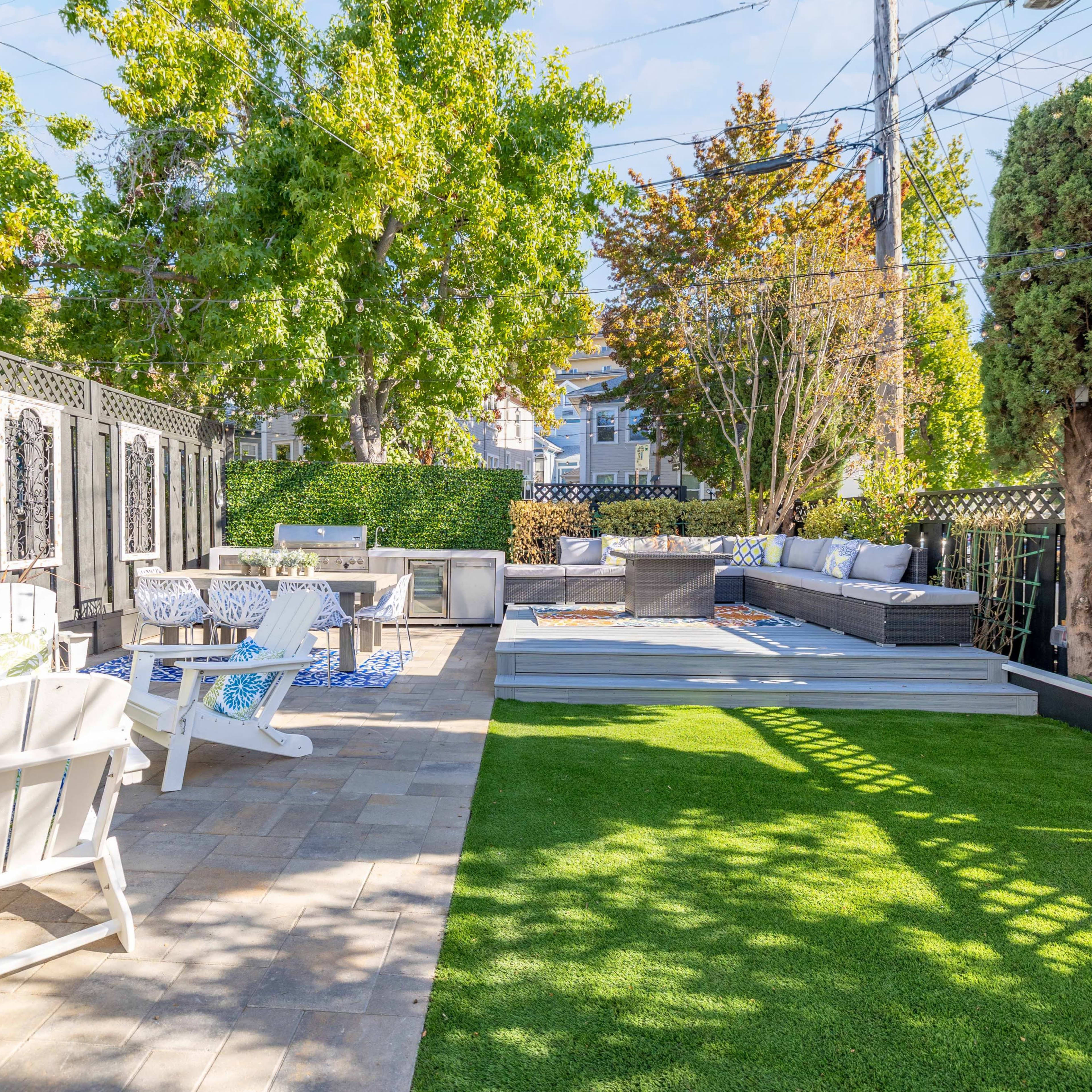 A landscaped backyard featuring a seating area with lounge furniture, a dining table, and a green lawn surrounded by trees and privacy screens.
