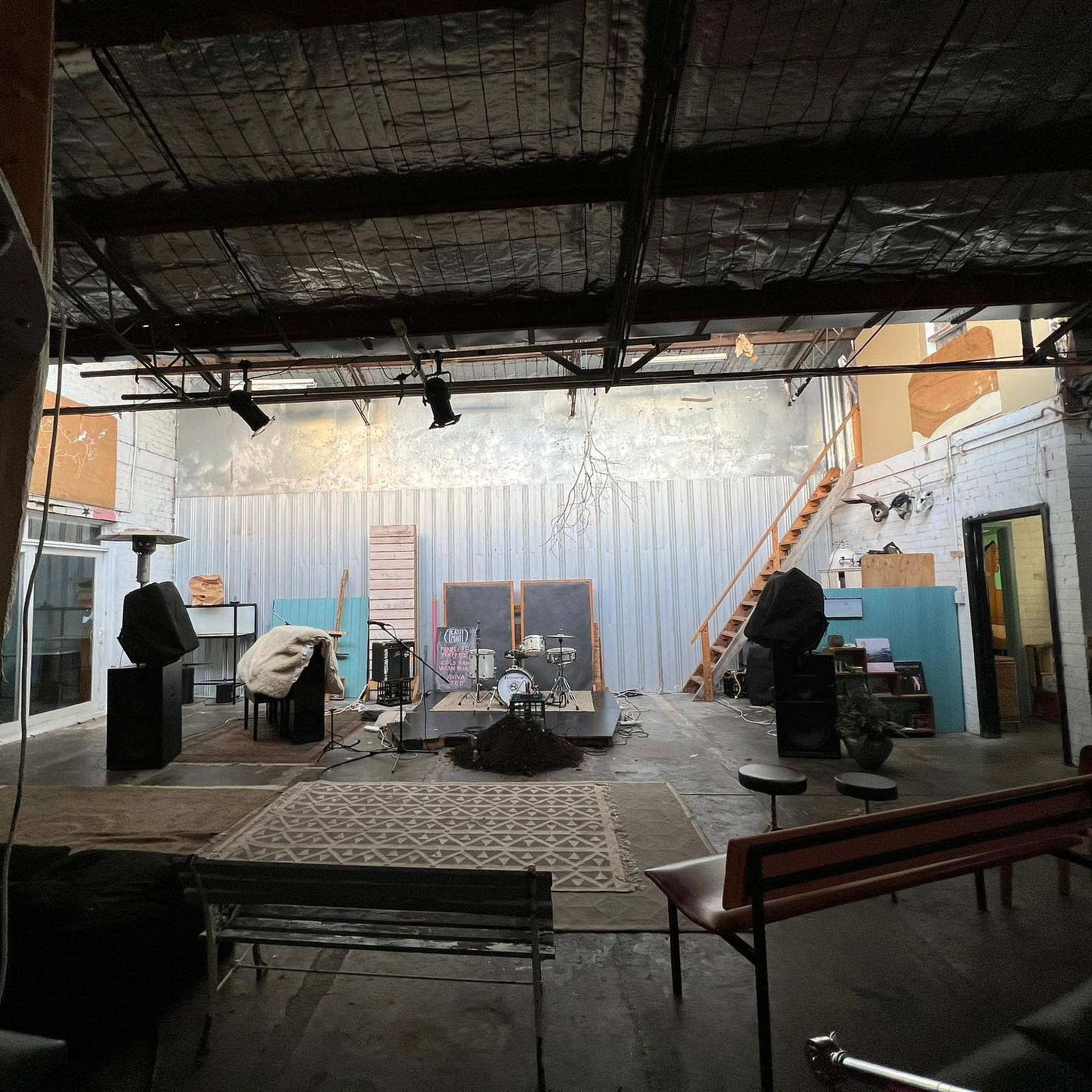 A spacious, empty indoor area with a wooden staircase, scattered musical equipment, and a few chairs arranged on a textured floor.