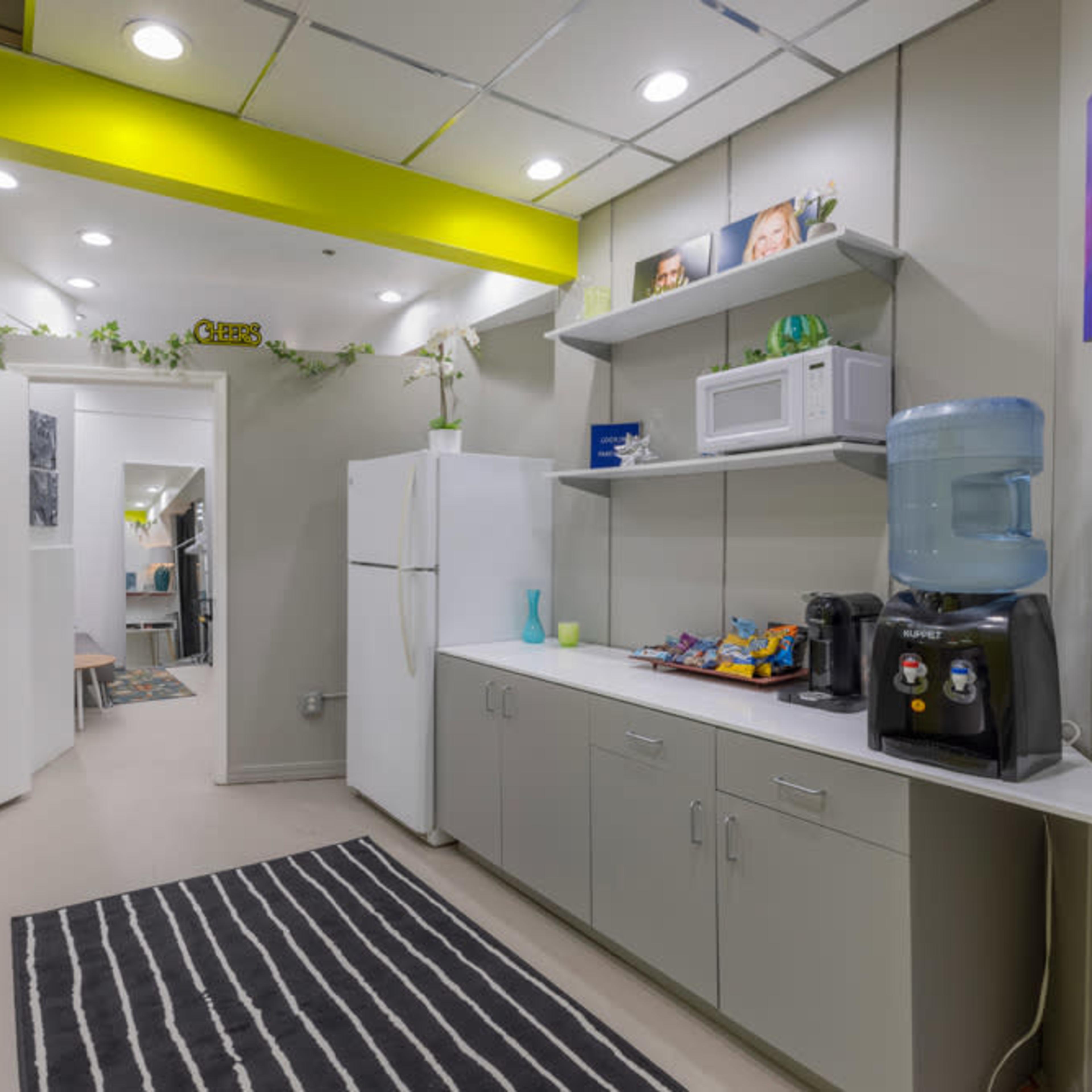 A modern break room with a kitchenette featuring a microwave, water cooler, snacks, and a refrigerator, alongside a hallway leading to another room.