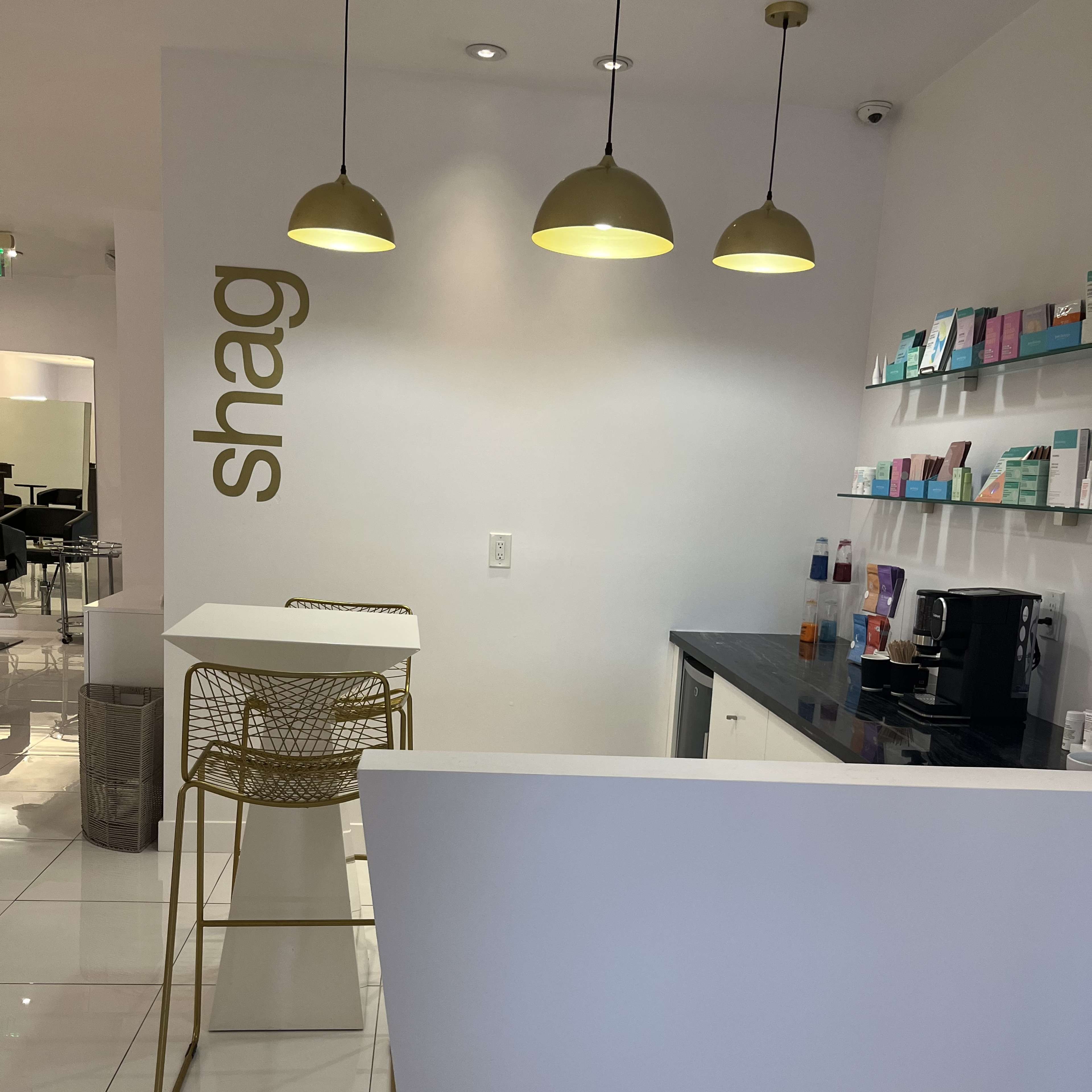 A modern reception area with a white wall, gold pendant lights, and a small counter with a coffee machine and product display shelves.