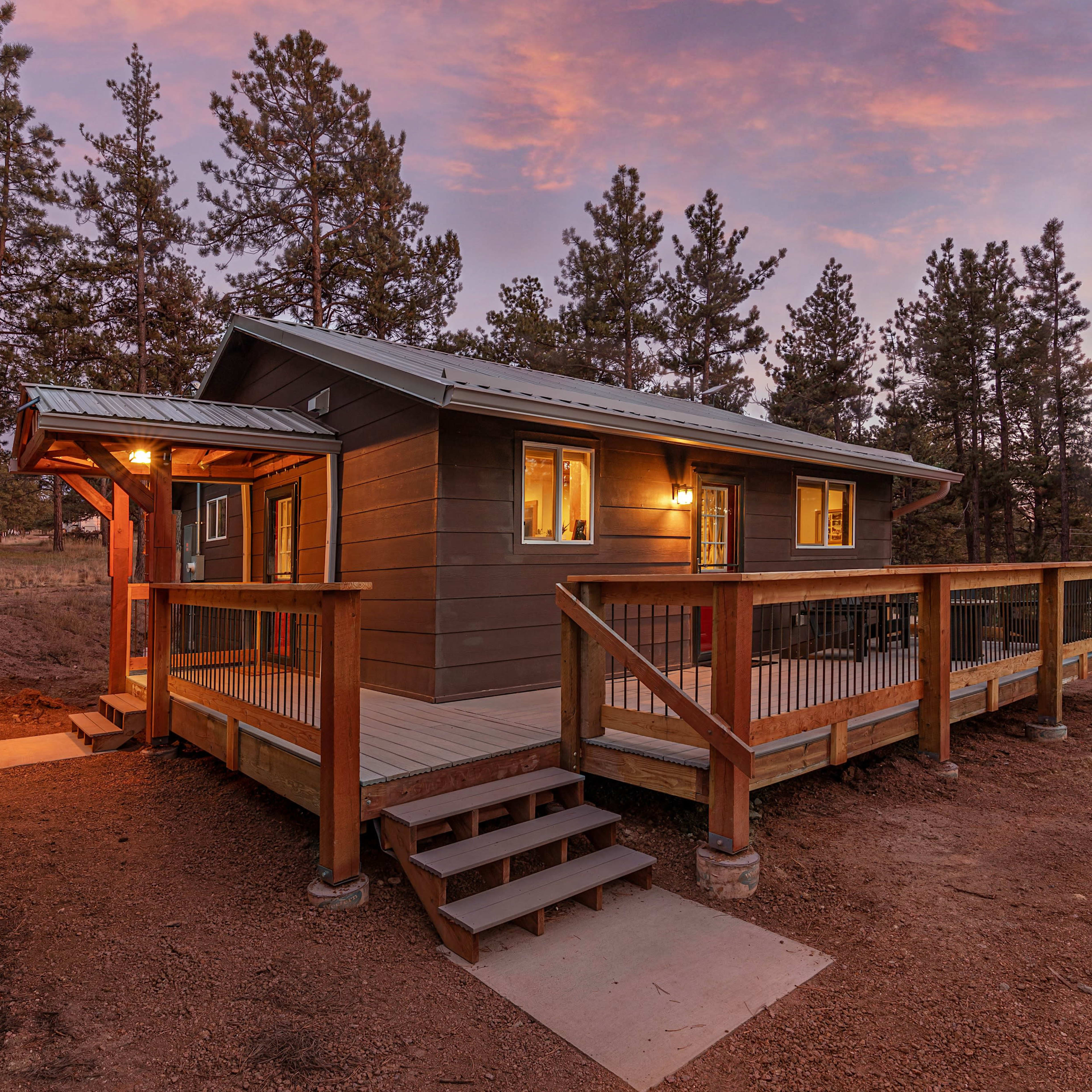 A wooden cabin with a deck and stairs is set among tall pine trees under a colorful sunset sky.