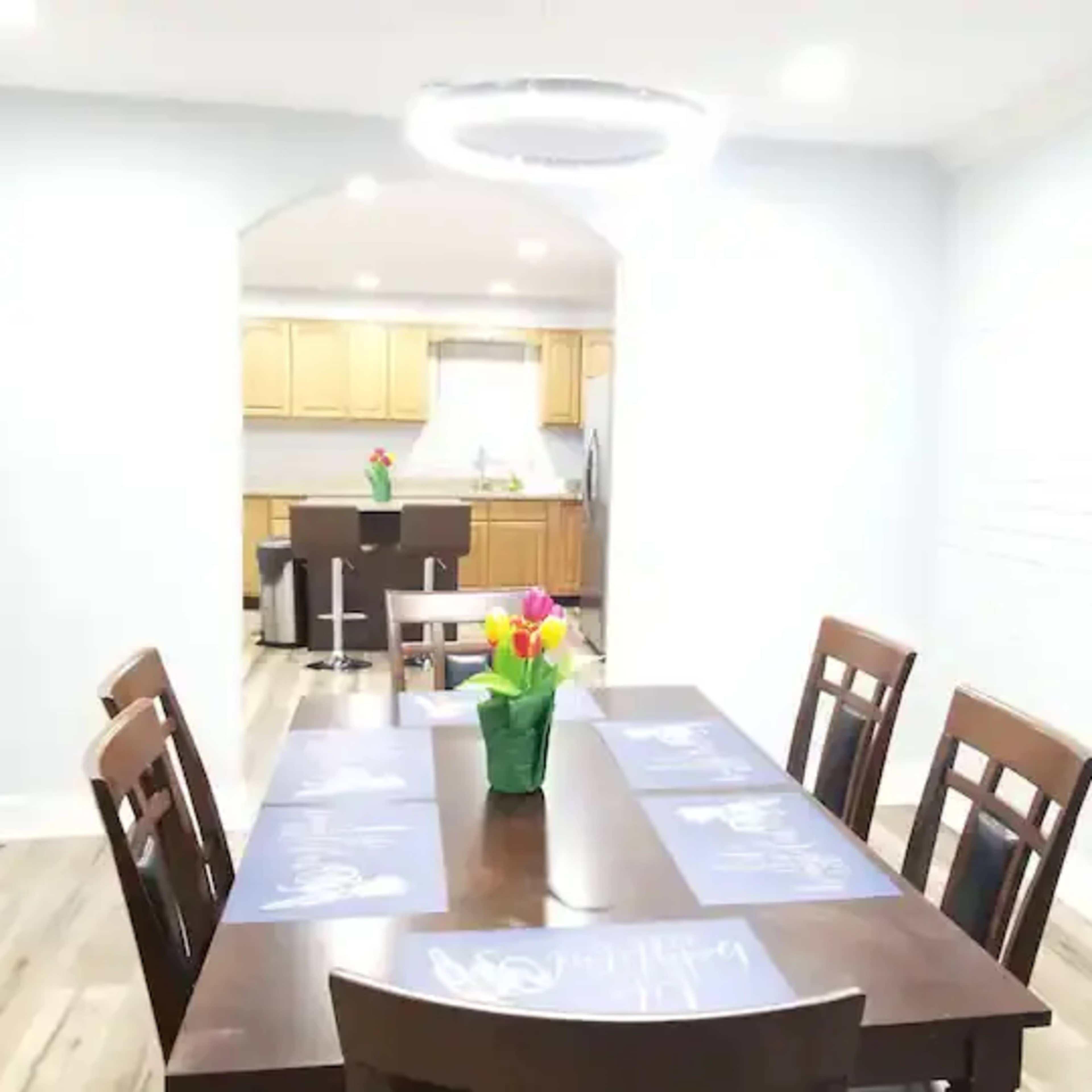 A dining area features a wooden table with a flower arrangement, surrounded by chairs, and a kitchen visible in the background.