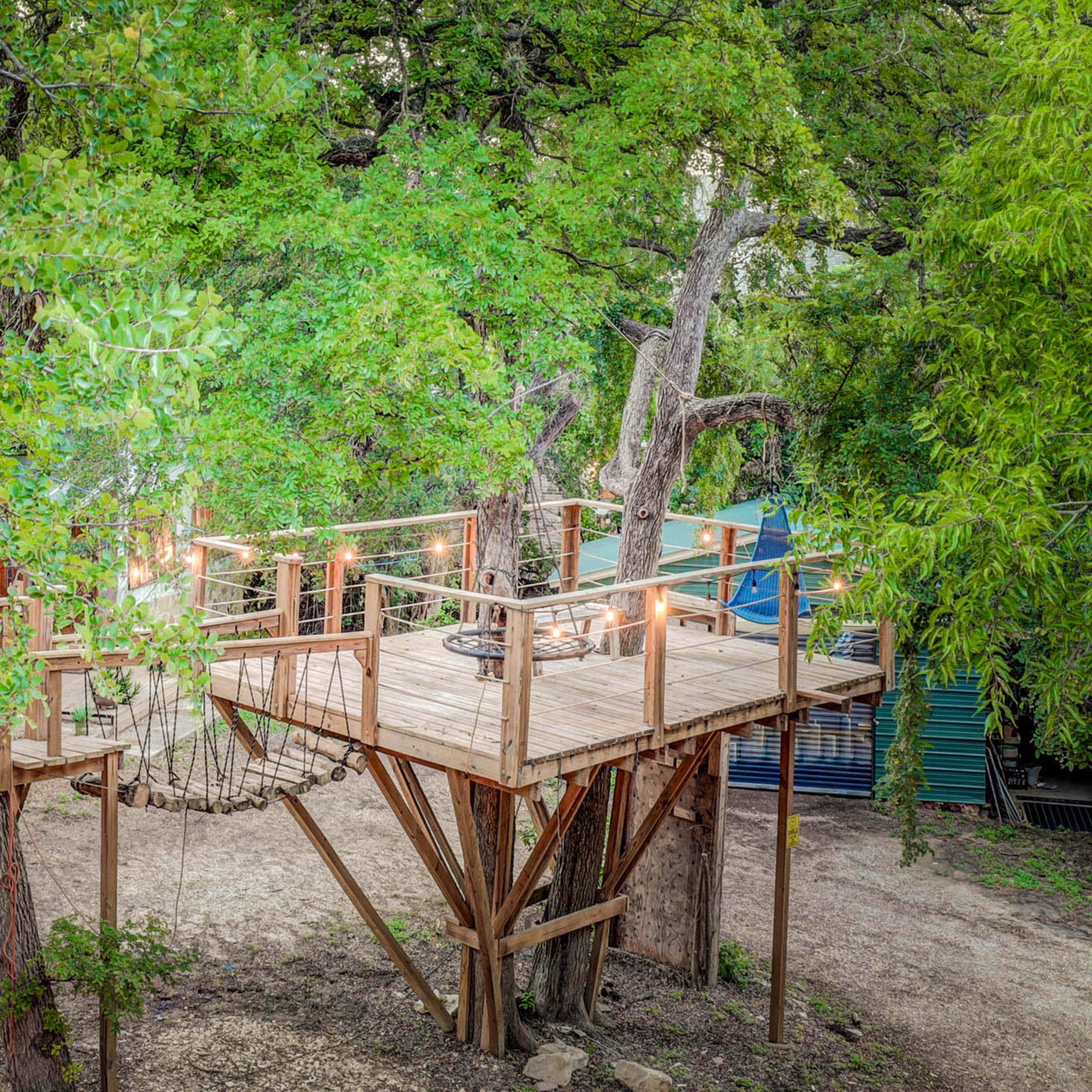 A wooden treehouse platform is elevated among green trees, connected by a rope bridge to another structure.