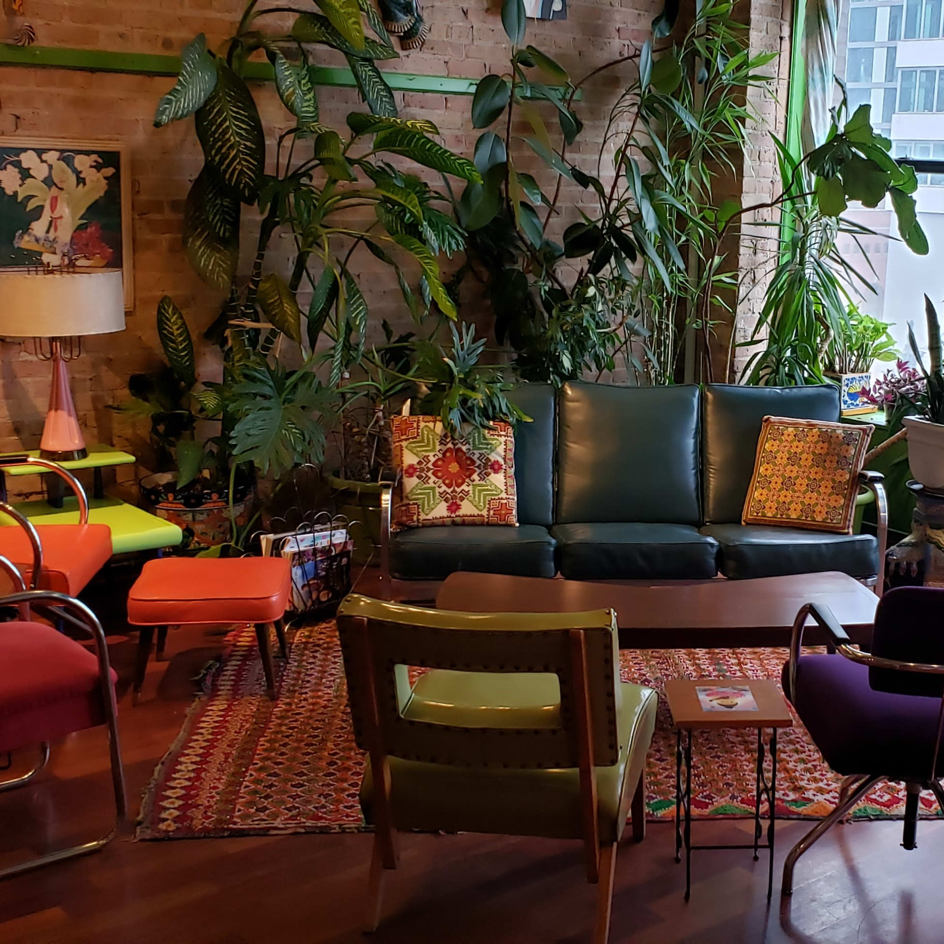 A cozy sitting area features a green couch, colorful chairs, a patterned rug, and various plants against a backdrop of large windows.
