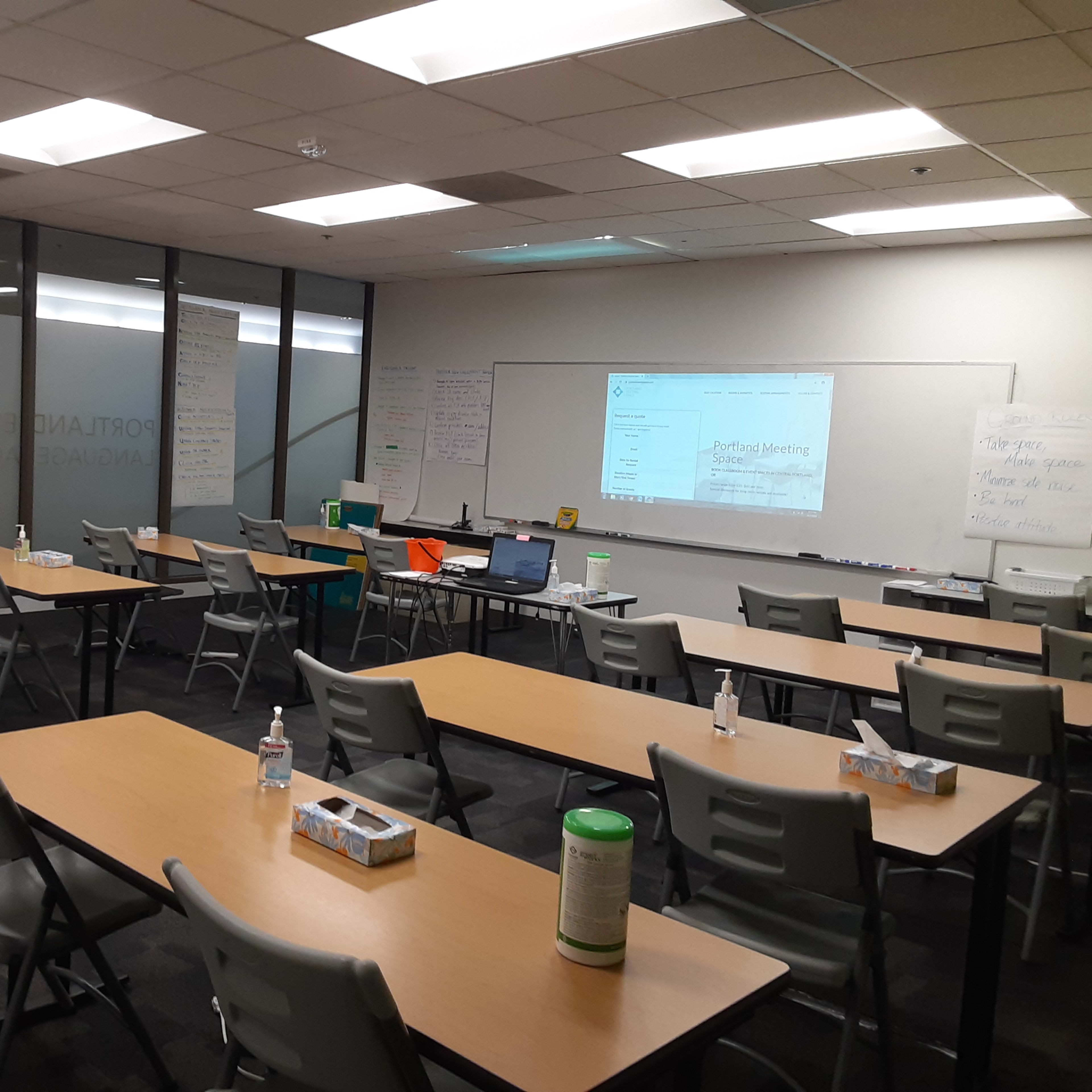 A classroom or training room with several tables and chairs arranged in rows, a projector displaying content on a screen, and whiteboards on the walls.