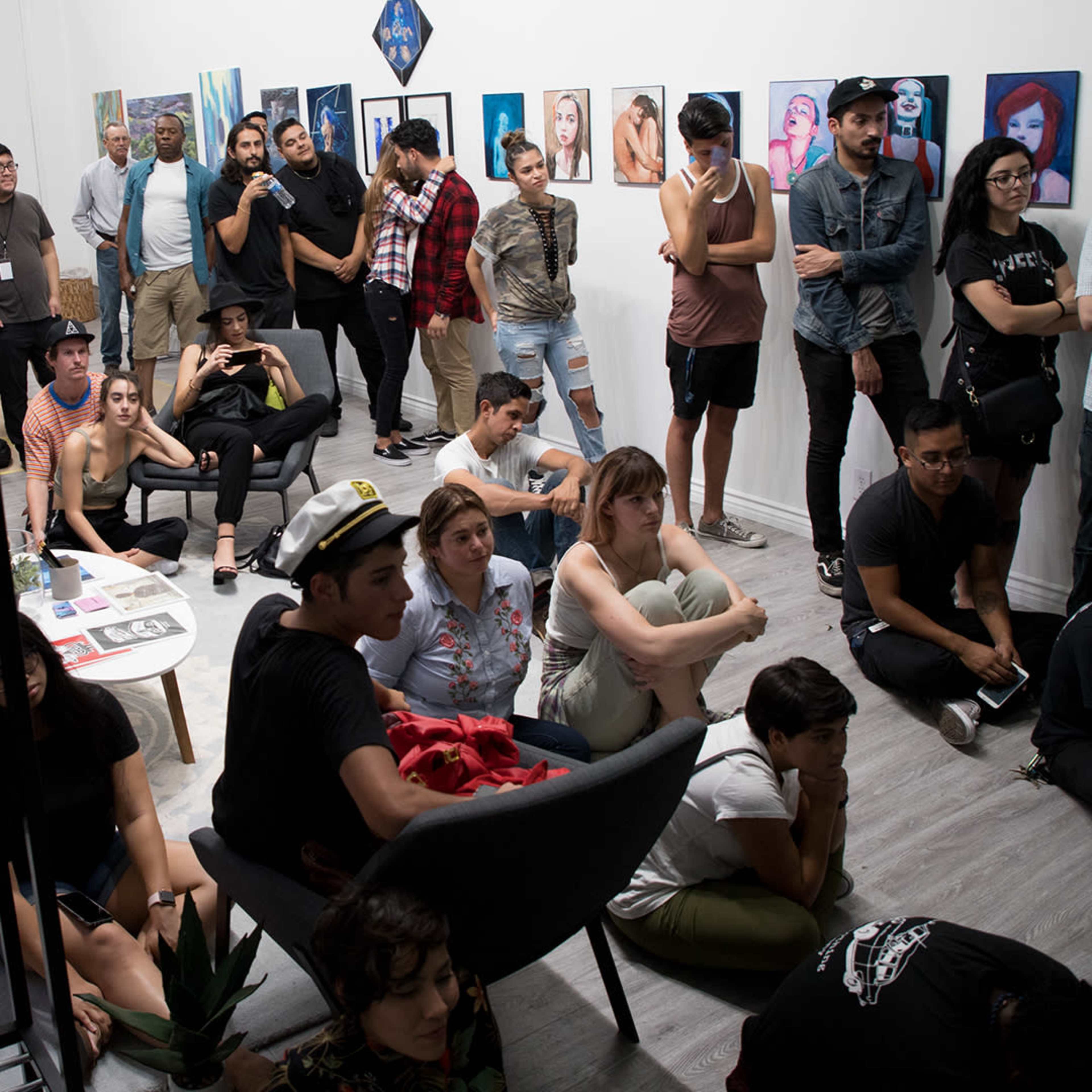 A diverse group of people is seated and standing in an art gallery, engaged in an event featuring artwork displayed on the walls.