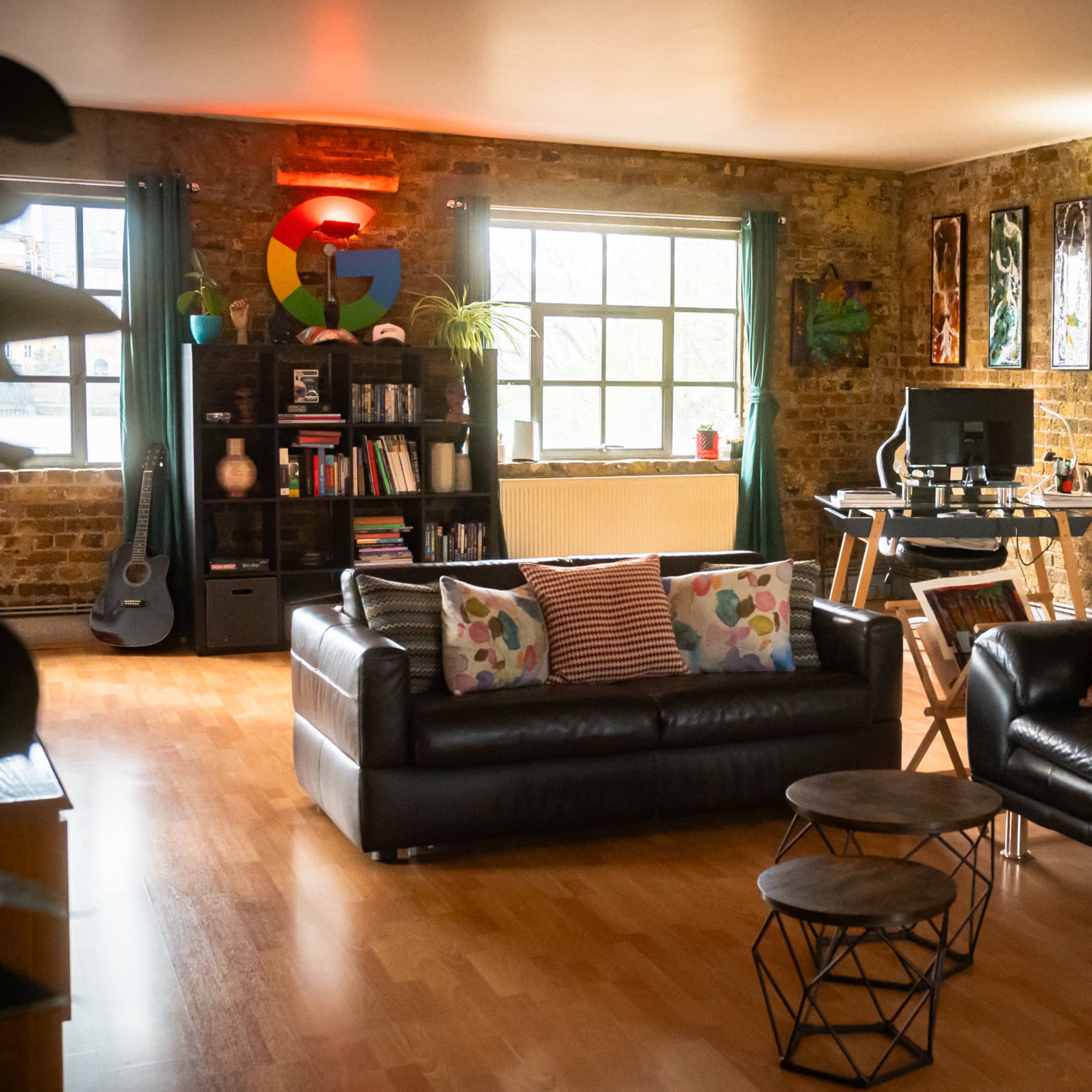 A cozy living room features two black sofas with colorful pillows, a wooden floor, a brick wall, and a small desk with a computer setup.