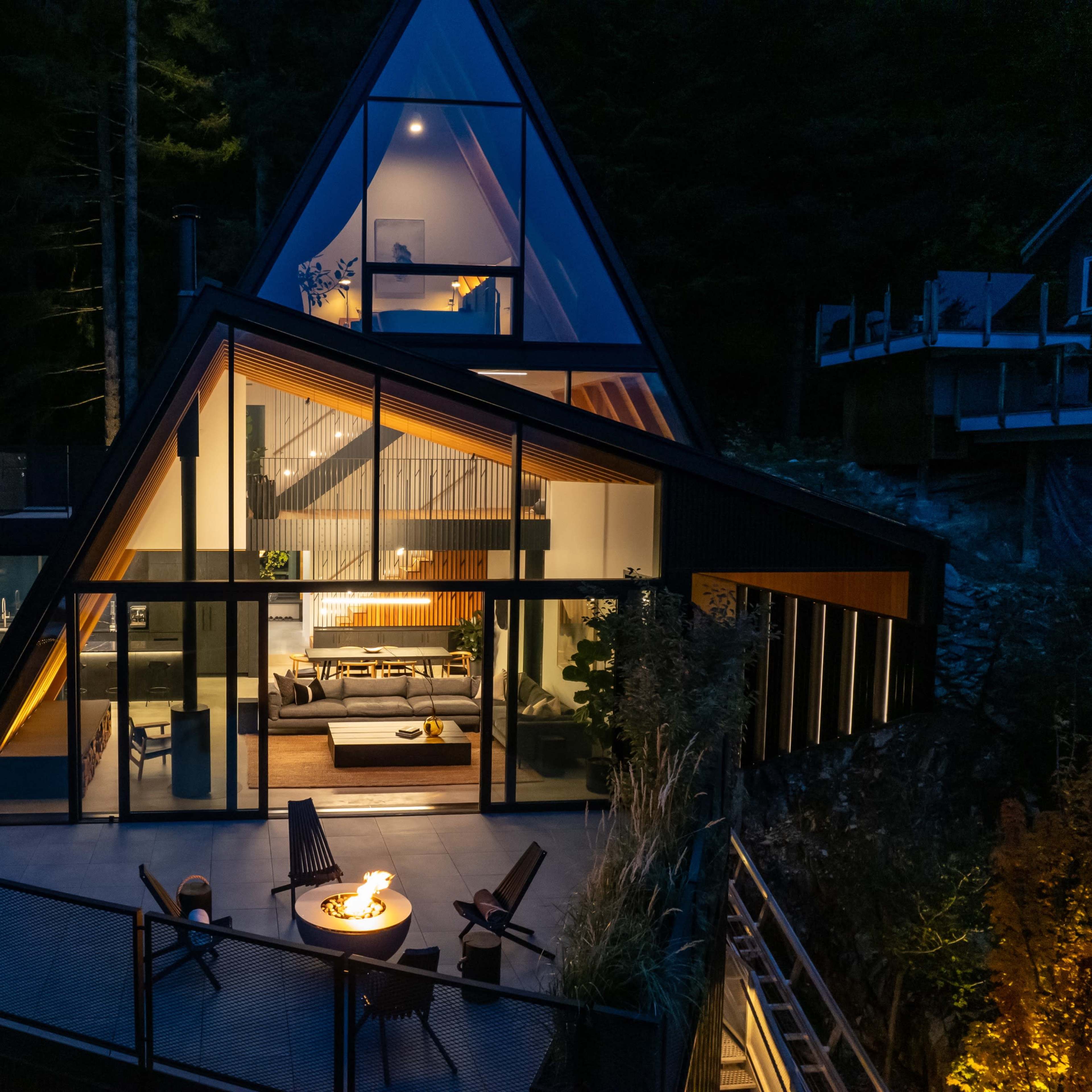 A modern A-frame house with large glass panels and a deck is illuminated by outdoor lighting at night, surrounded by trees.