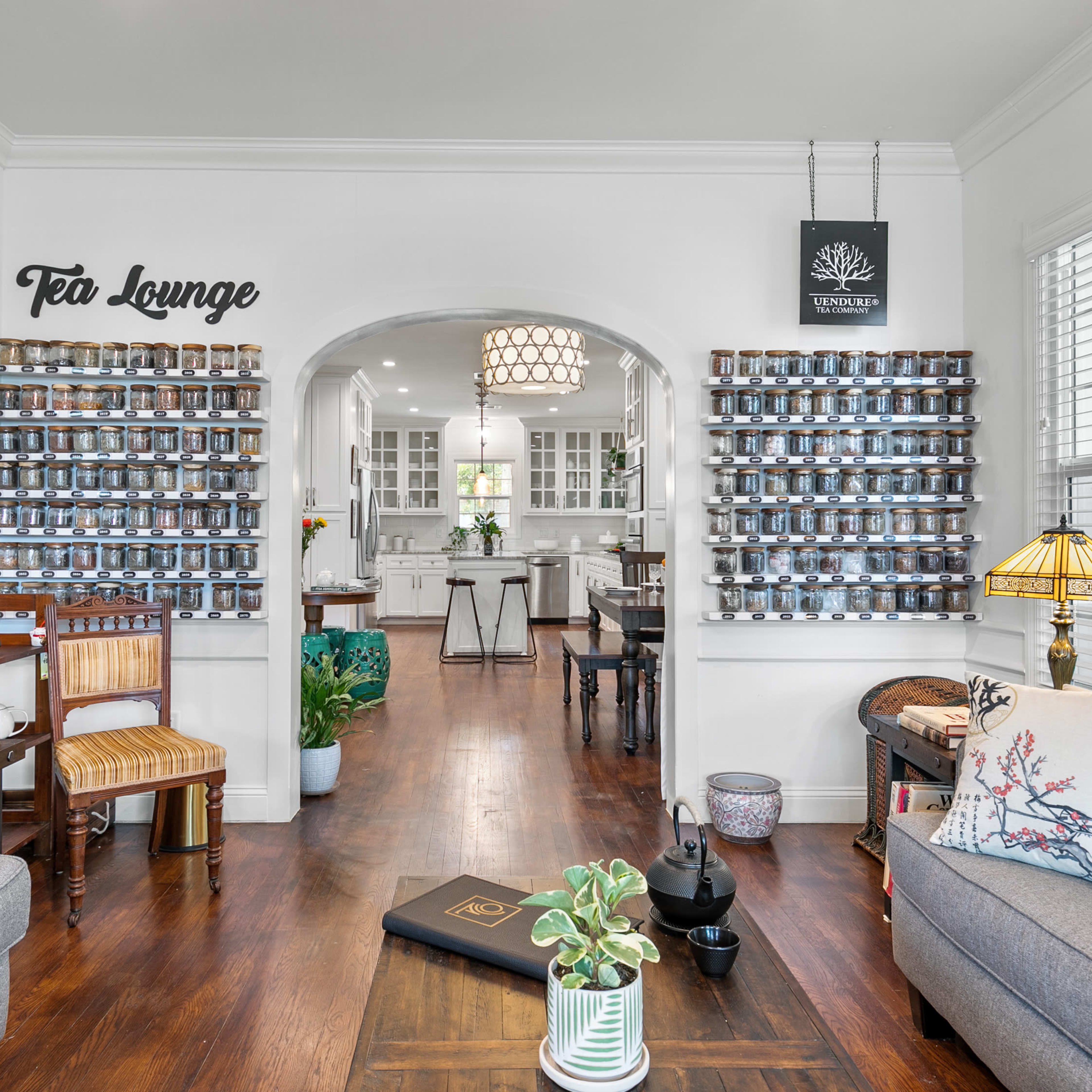 A cozy living area leading into a bright kitchen, featuring a tea lounge sign, a wall of jars, and comfortable seating.