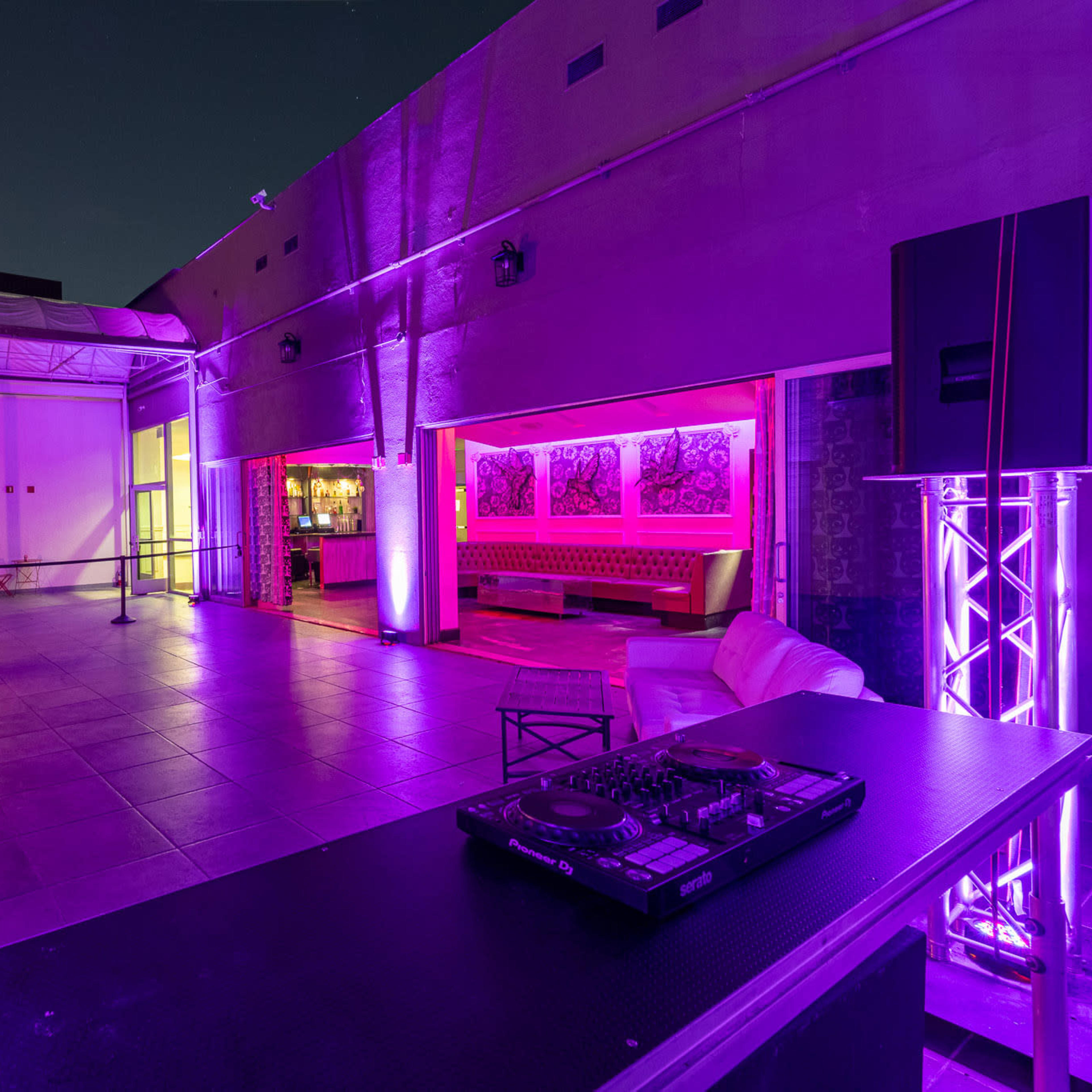 A modern outdoor lounge area with a DJ setup, illuminated by purple lights, and featuring a bar in the background.