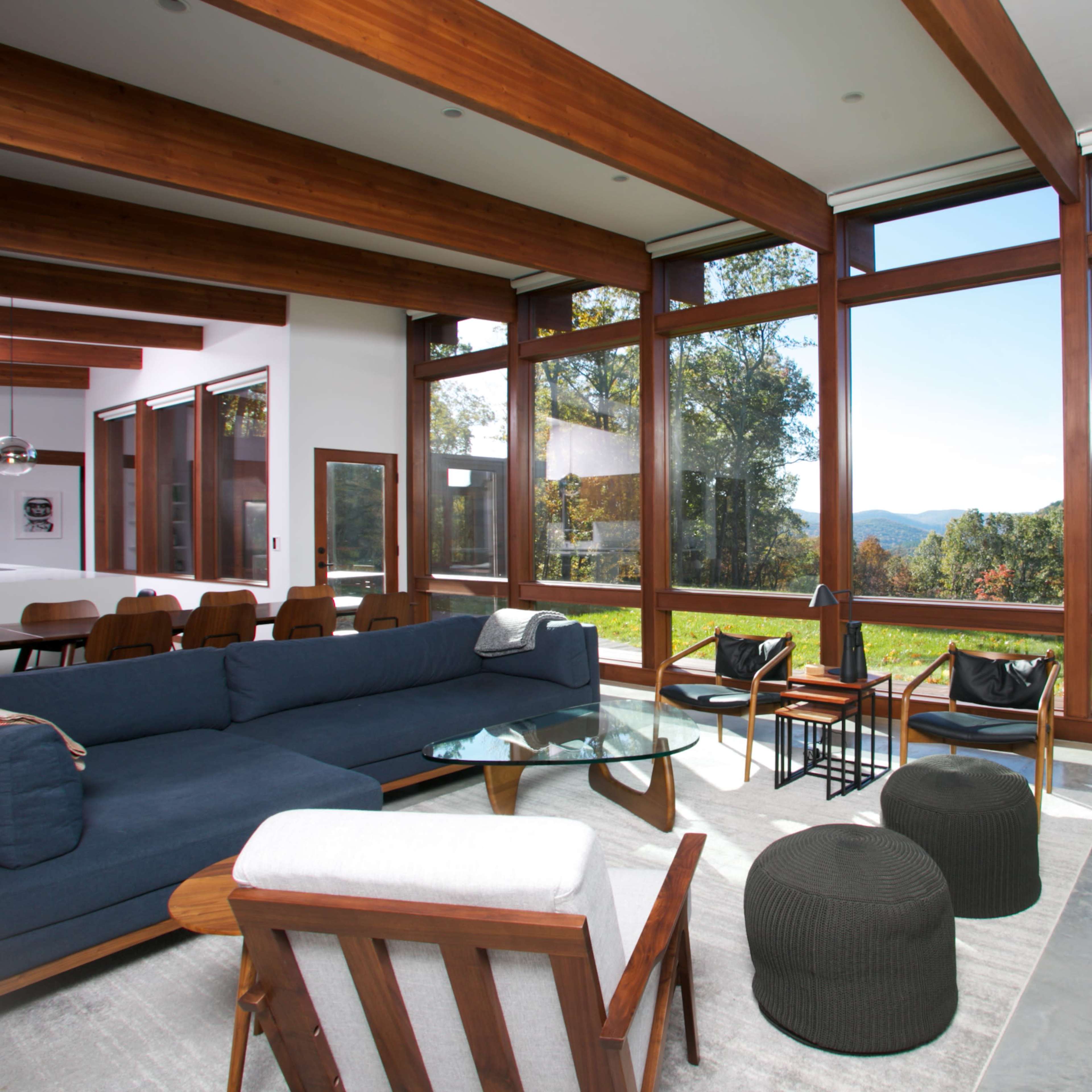 The scene features a modern living room with large windows offering a view of the landscape, furnished with a blue sofa, glass coffee table, and black accent pieces.