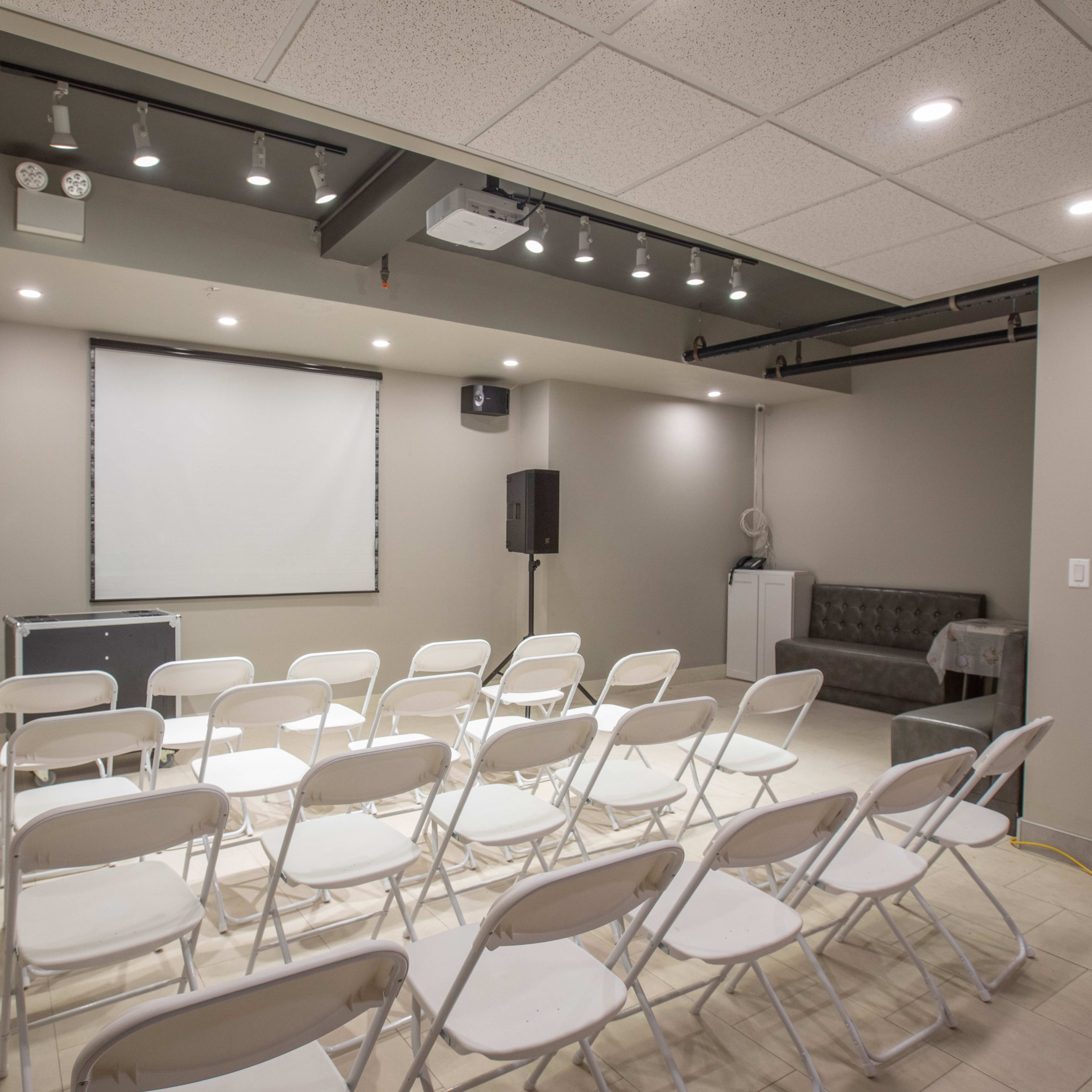 A seminar room with rows of white chairs facing a blank projection screen and a sound system in the corner.
