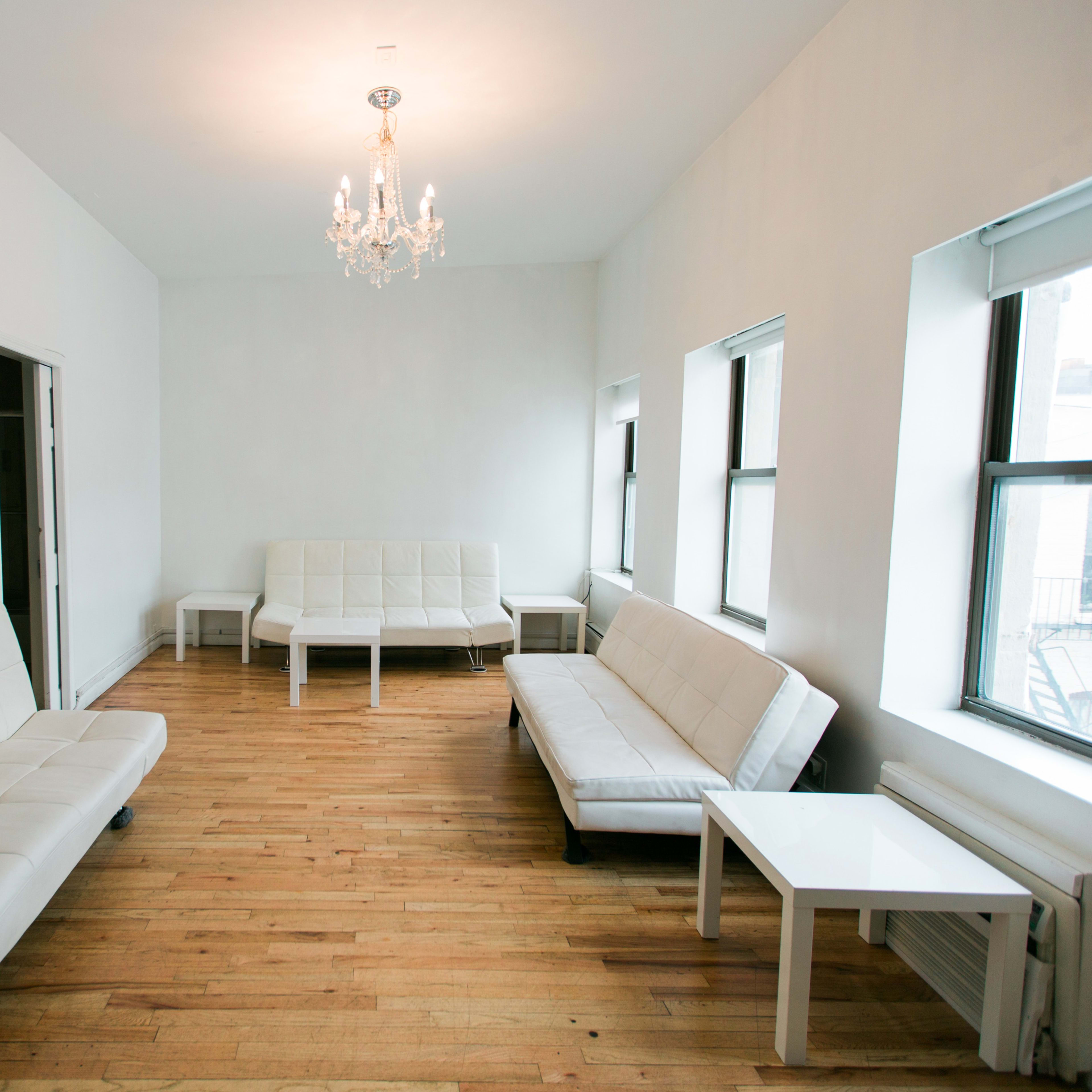 The room features white sofas, a white chandelier, and large windows on two walls, all set against a light-colored wood floor.