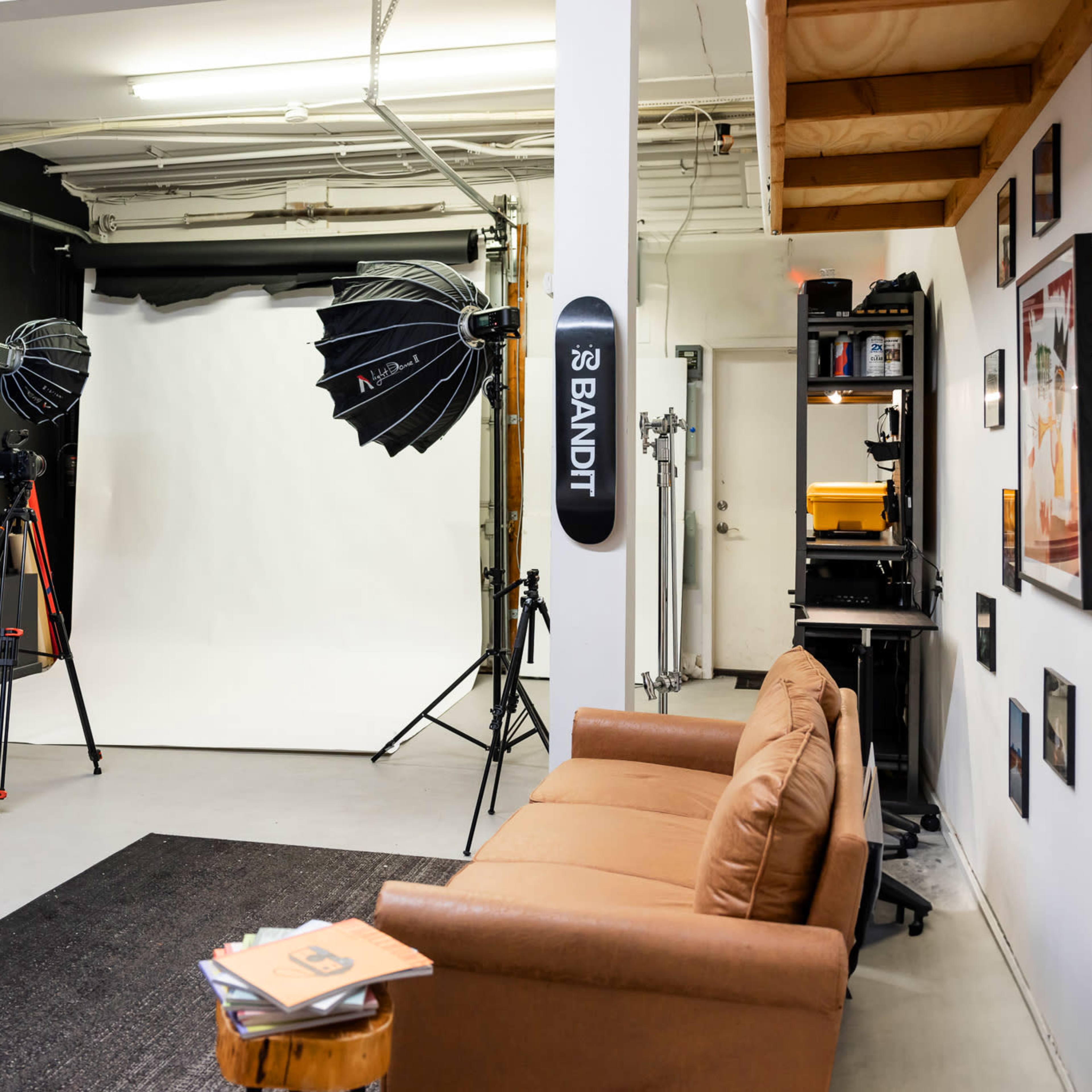 A photography studio with a black wall, a white backdrop, two softbox lights on tripods, a brown couch, and a shelf against the wall filled with equipment.