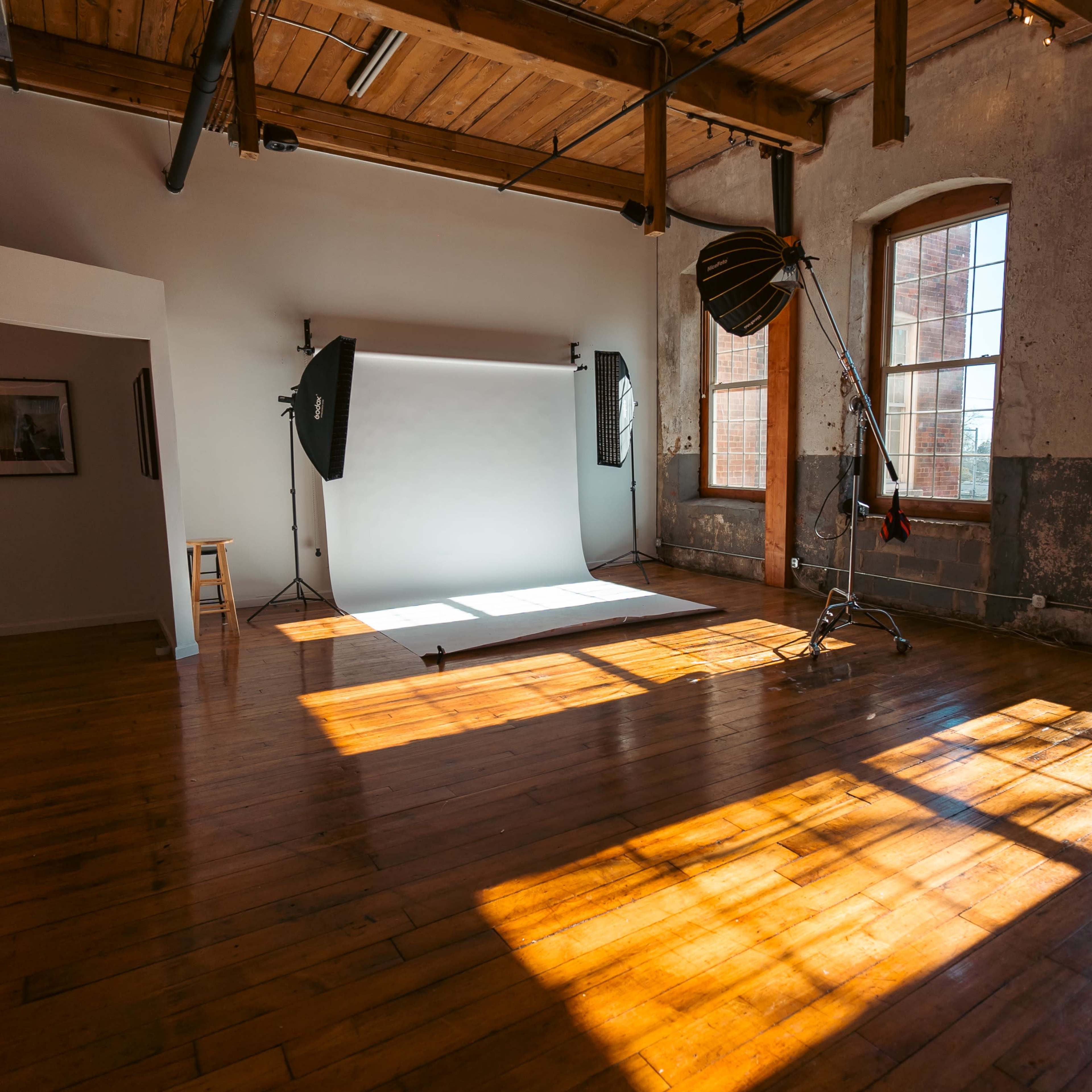 A spacious photography studio with large windows, wooden floors, and a backdrop setup on one side.