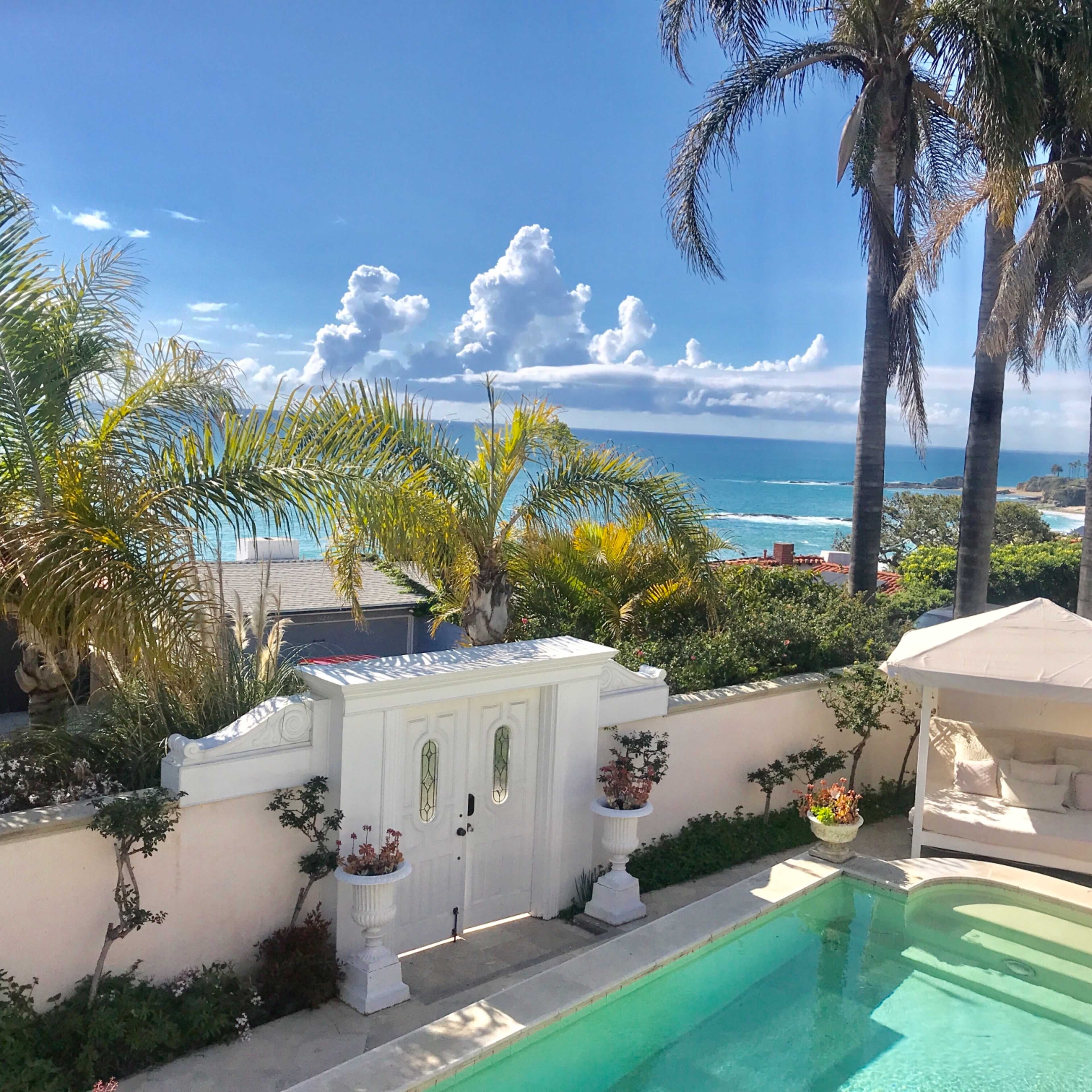 A view of a swimming pool surrounded by tropical plants, with the ocean and clouds visible in the background.