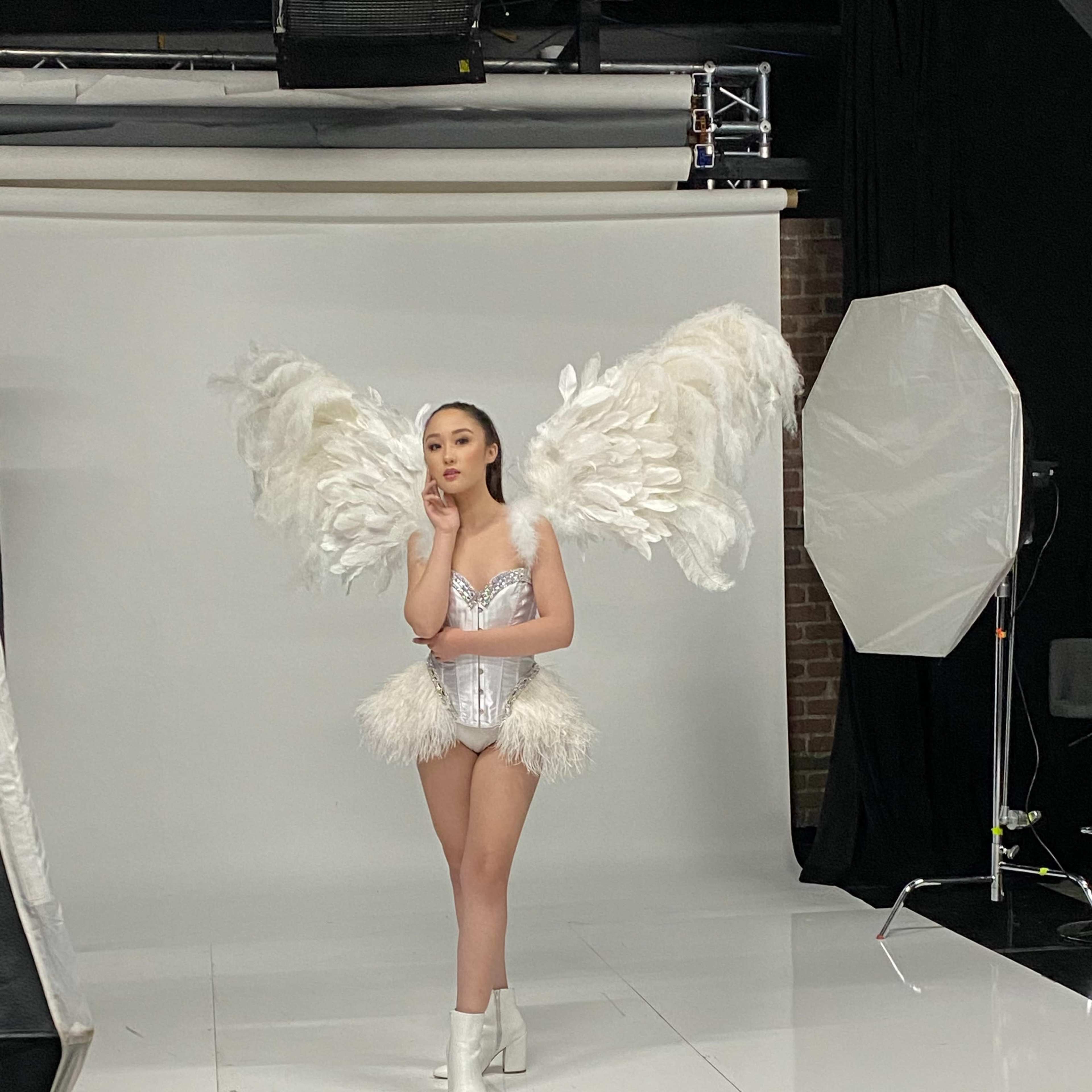 A model poses in a photoshoot wearing a feathered costume with large wings and shiny white boots.