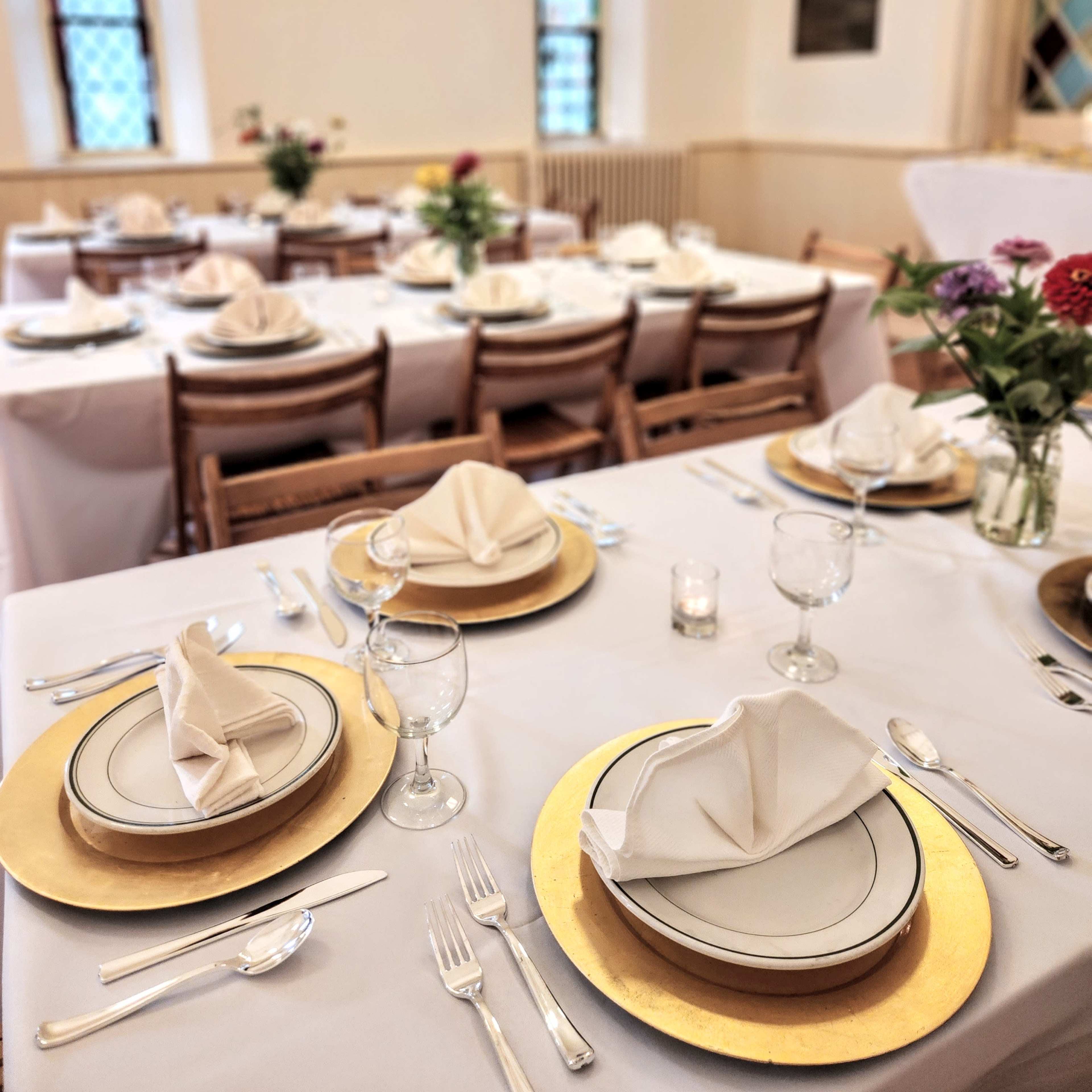 A set dining arrangement featuring white tablecloths, gold chargers, and neatly folded napkins in an elegantly decorated room with large windows.
