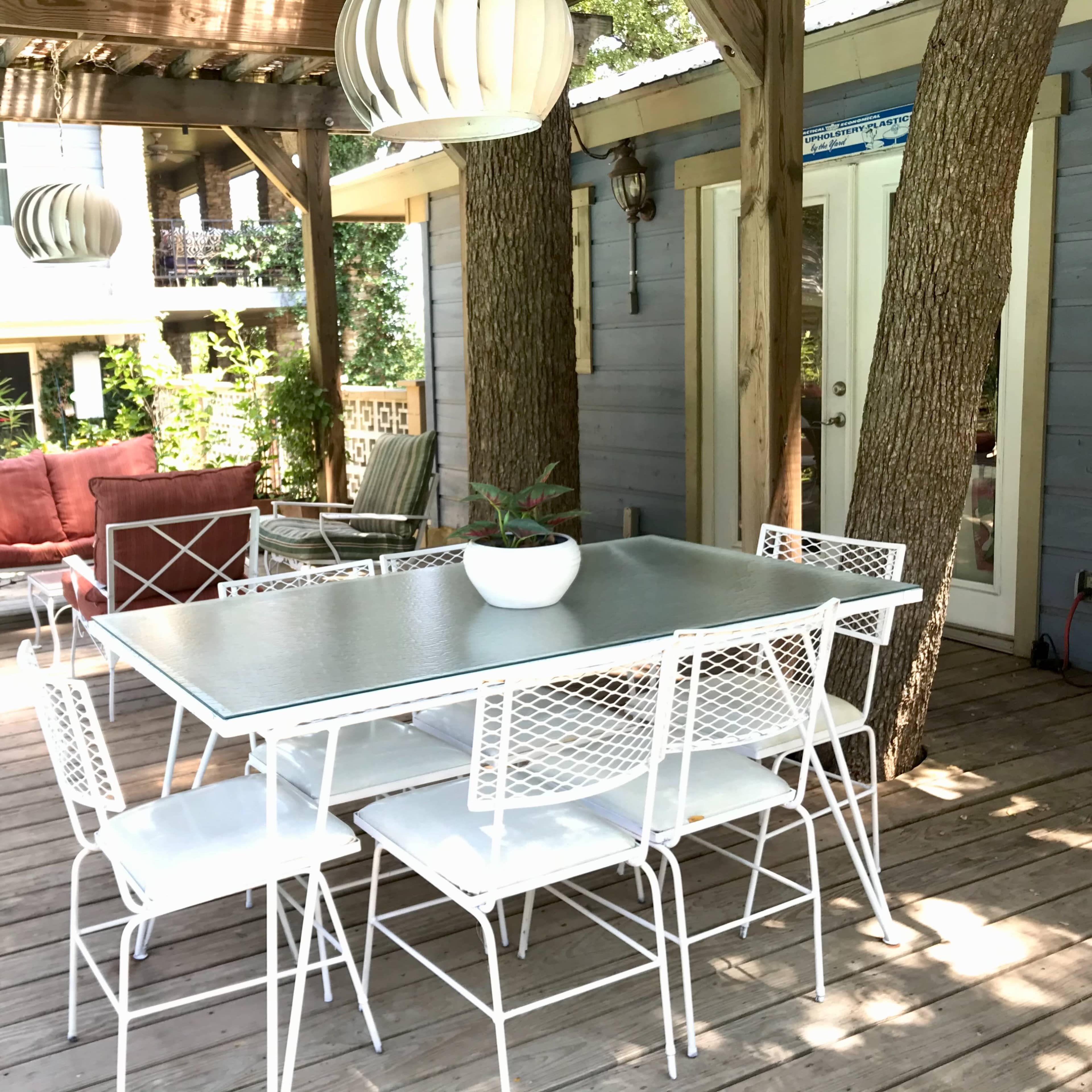 A patio area with a glass-top dining table surrounded by white metal chairs, complemented by potted plants and a seating arrangement nearby.