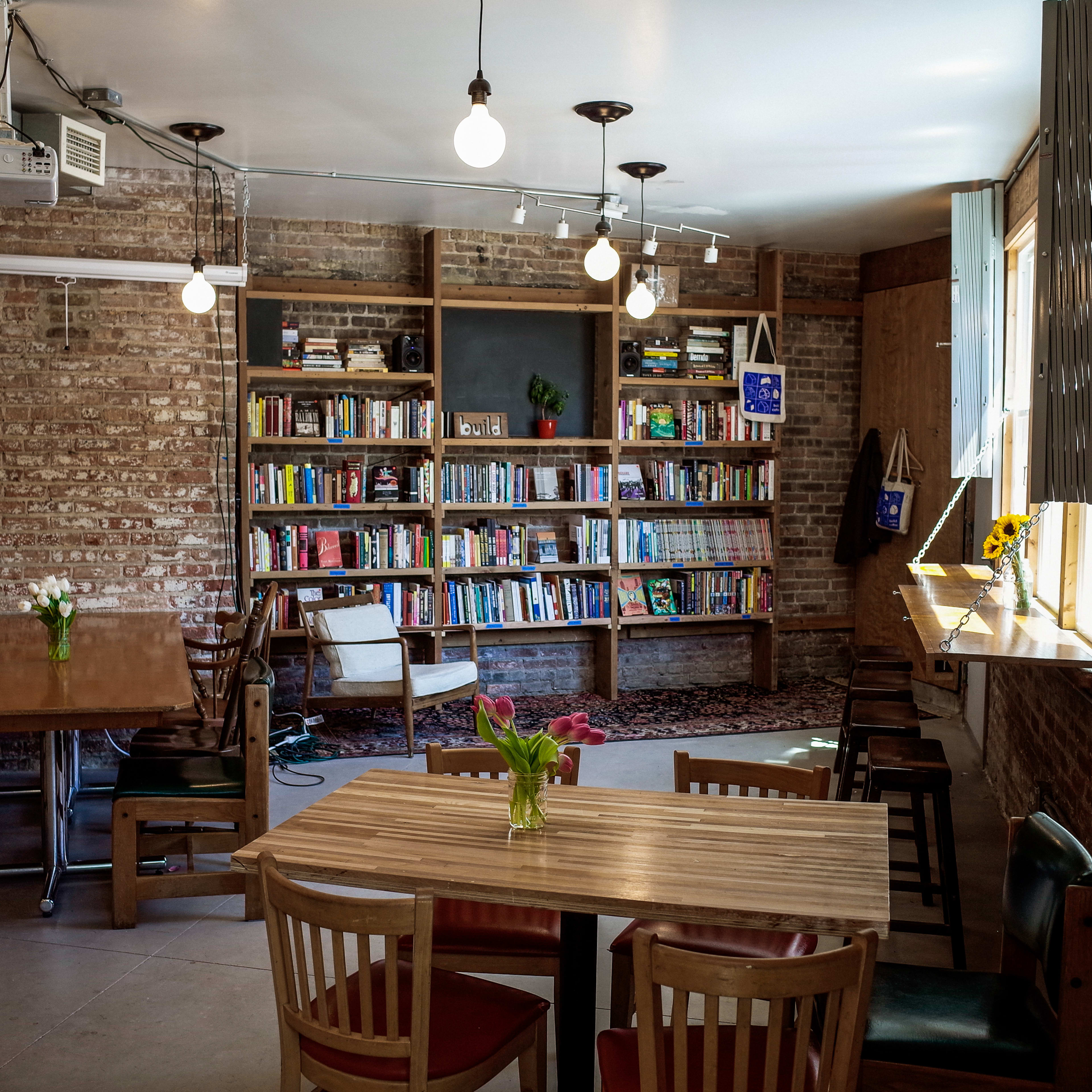 The scene features a cozy café with wooden tables and chairs, illuminated by hanging light bulbs, and a bookshelf filled with books against a brick wall.
