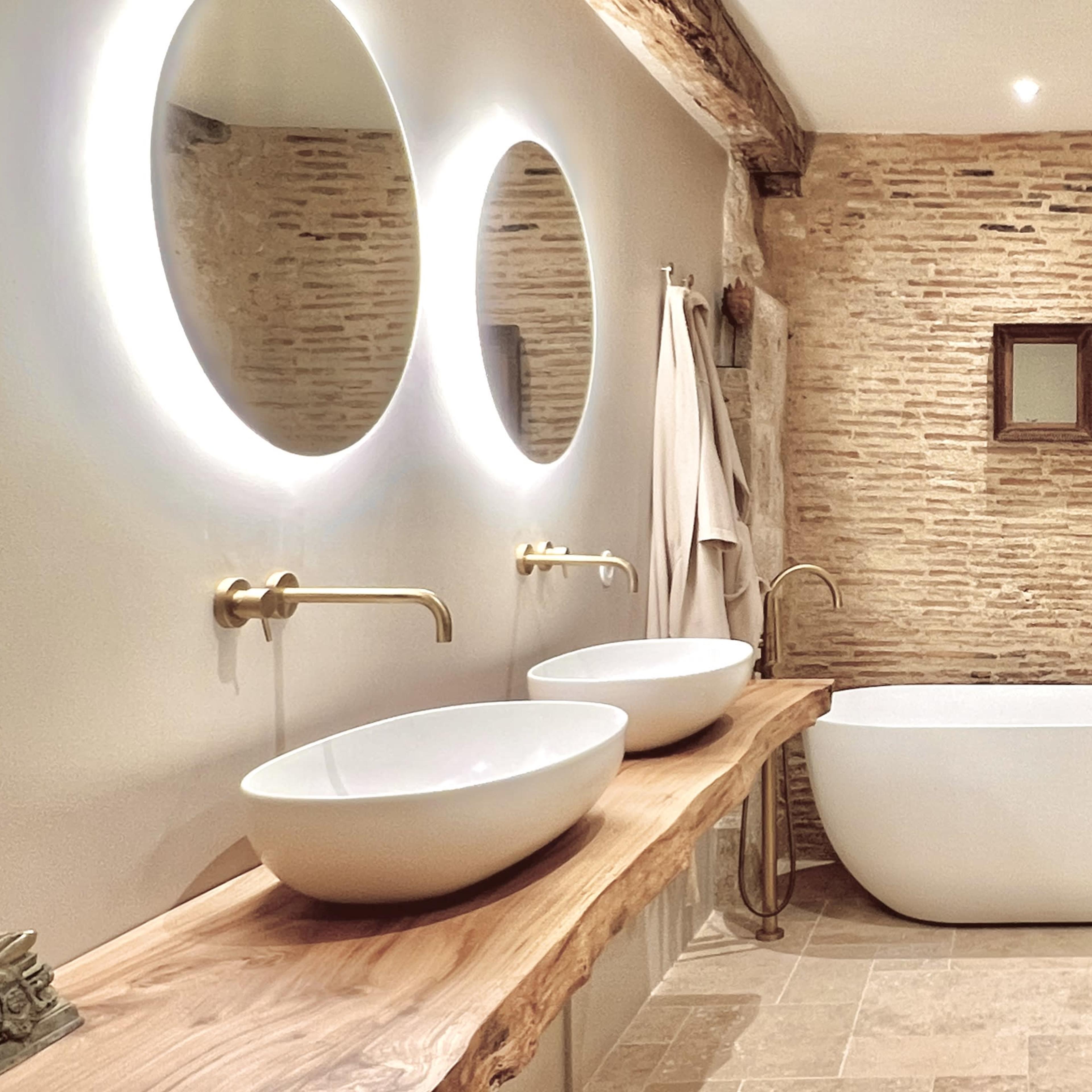 A modern bathroom featuring a freestanding white bathtub, two round sinks on a wooden countertop, and textured stone walls.