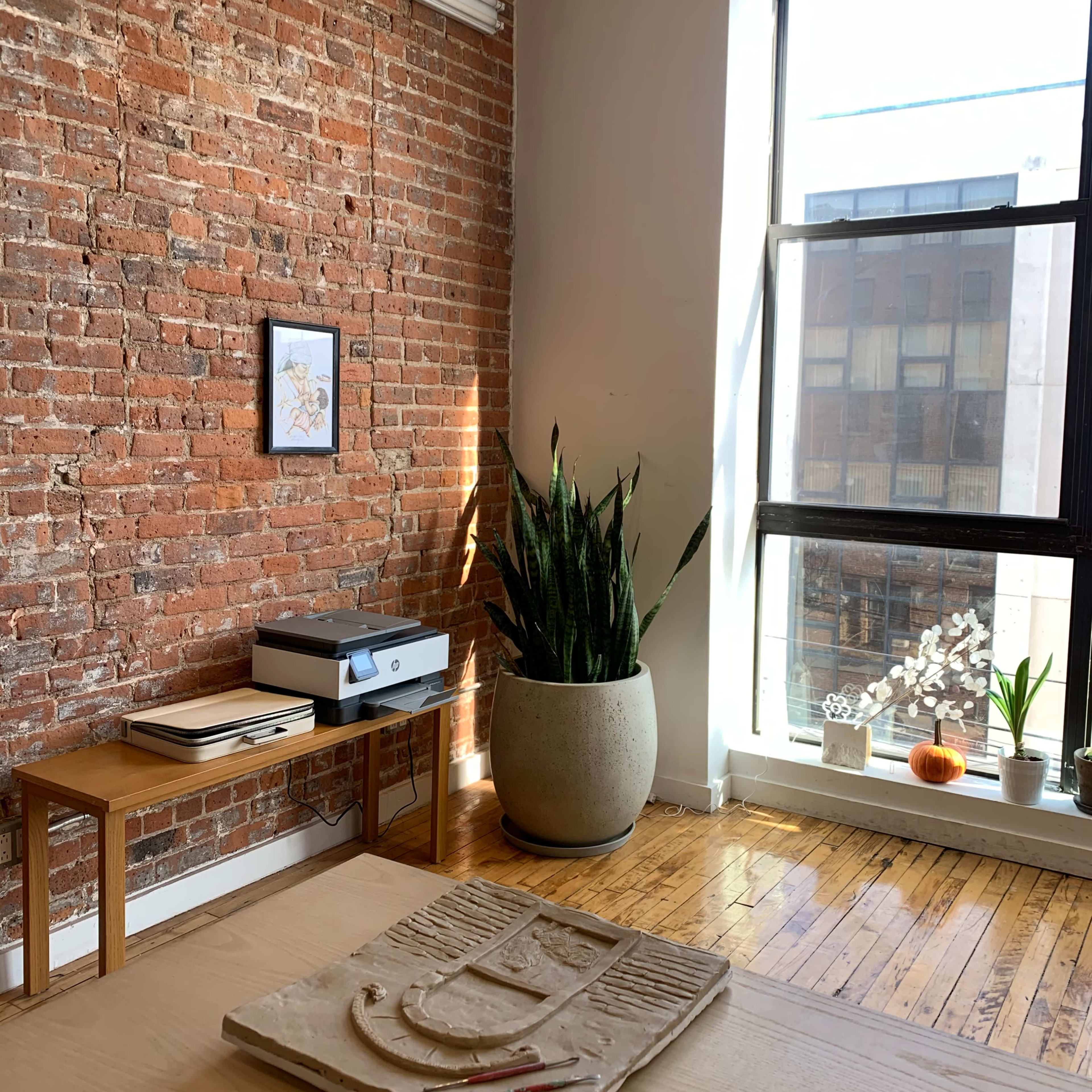 A minimalist workspace with a wooden table at the center, a brick wall, plants in the corner, and a window letting in natural light.