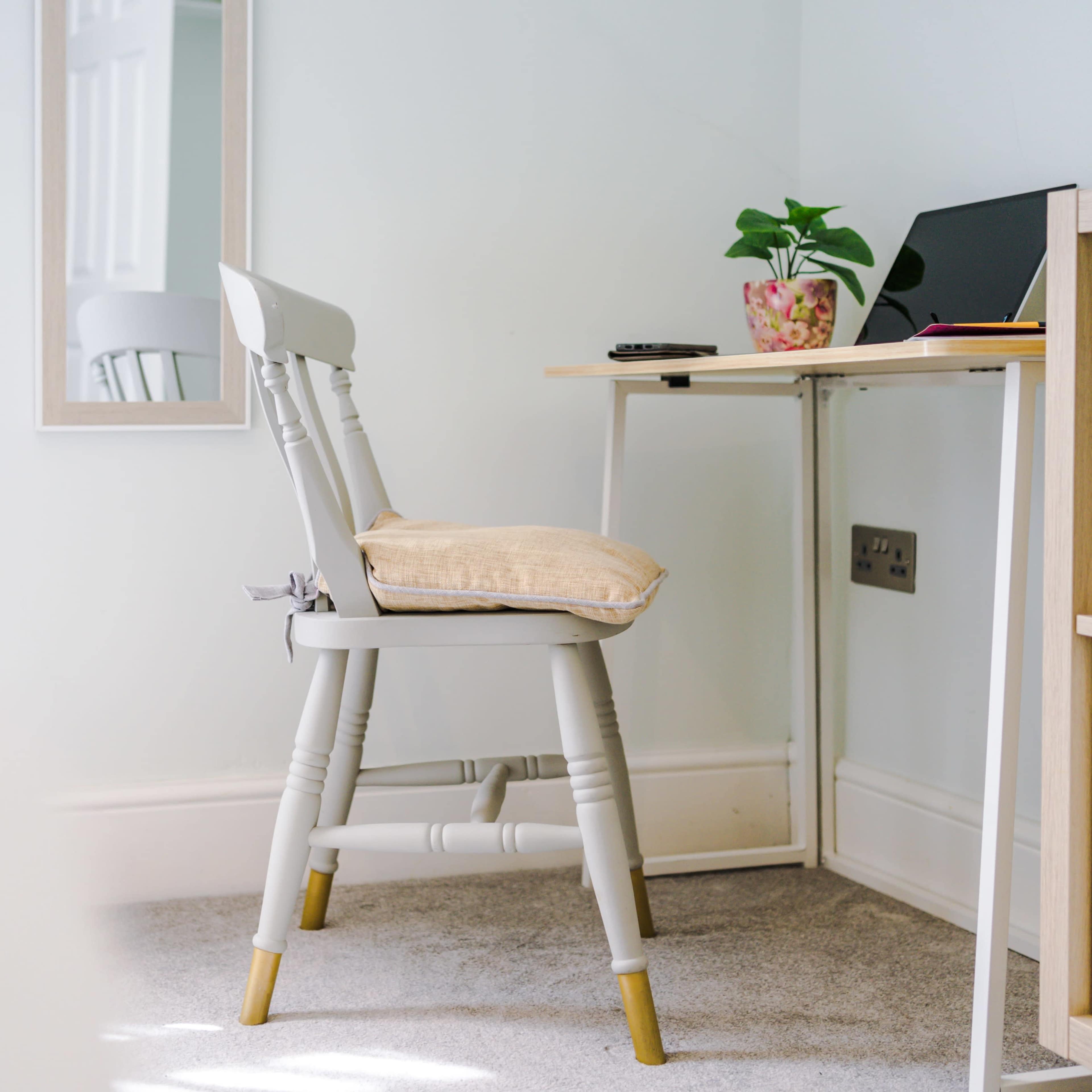 A light-colored chair with a cushion sits next to a small desk that has a laptop and a potted plant on it, in a room with pale blue walls.