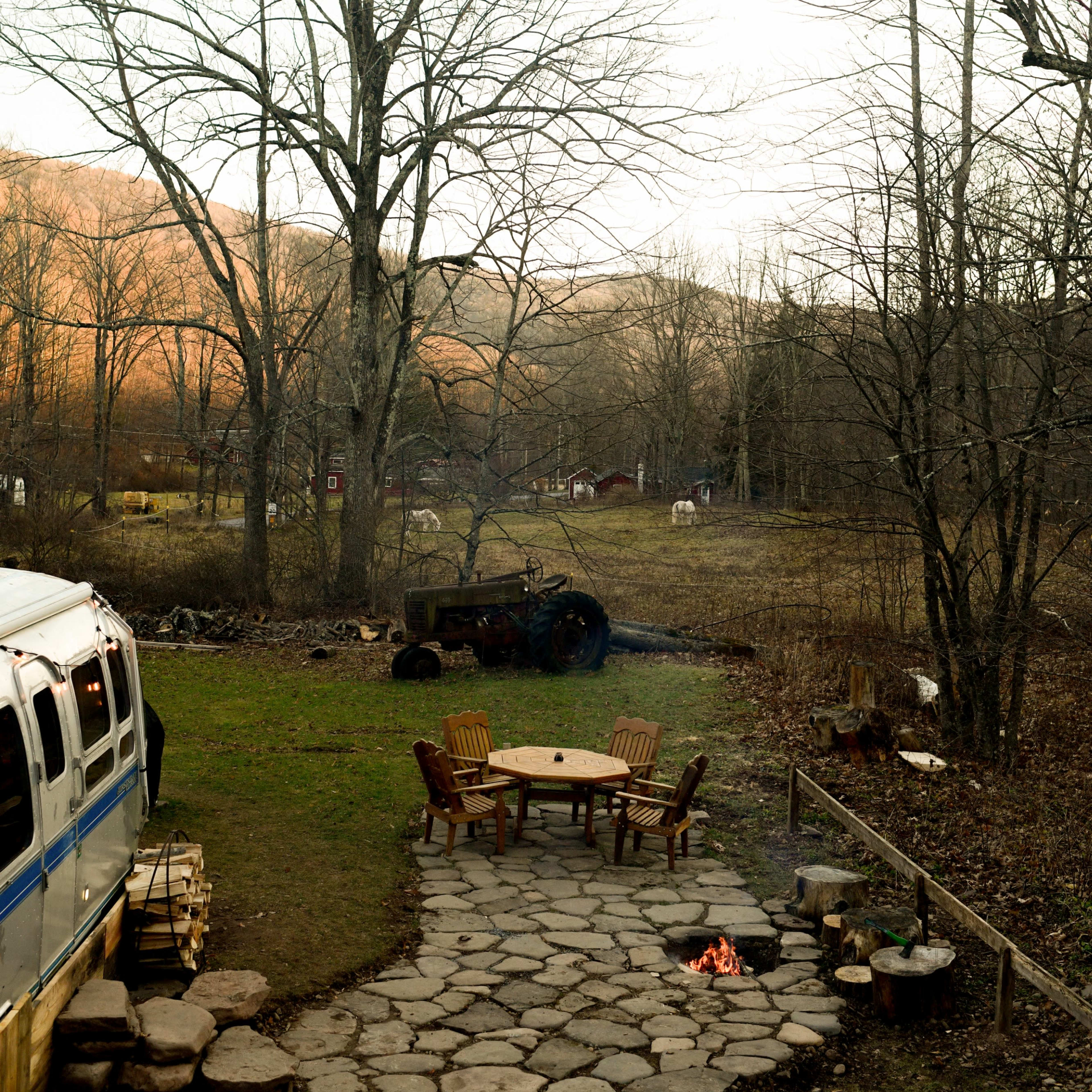 A stone patio with chairs surrounds a small fire pit, and an old truck is parked in a rural field with trees and a van in the foreground.