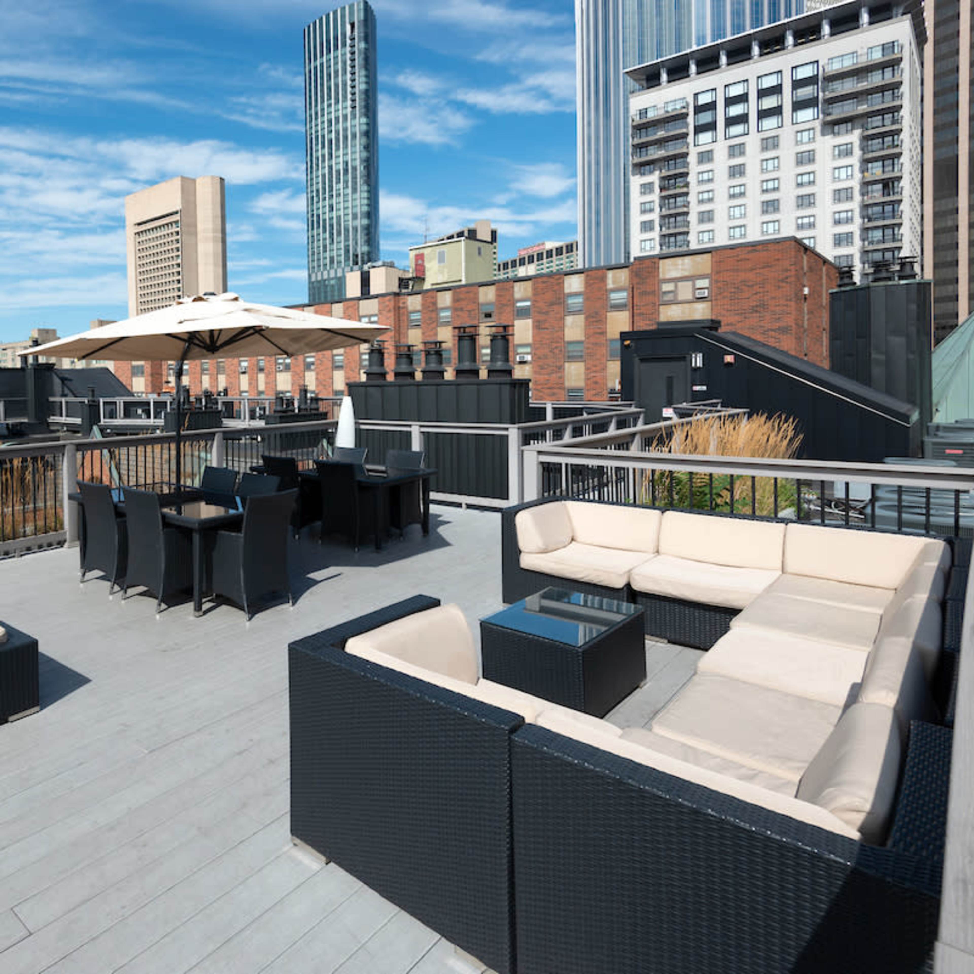 A rooftop terrace features outdoor seating arrangements, a dining area, and a large umbrella, surrounded by urban buildings.