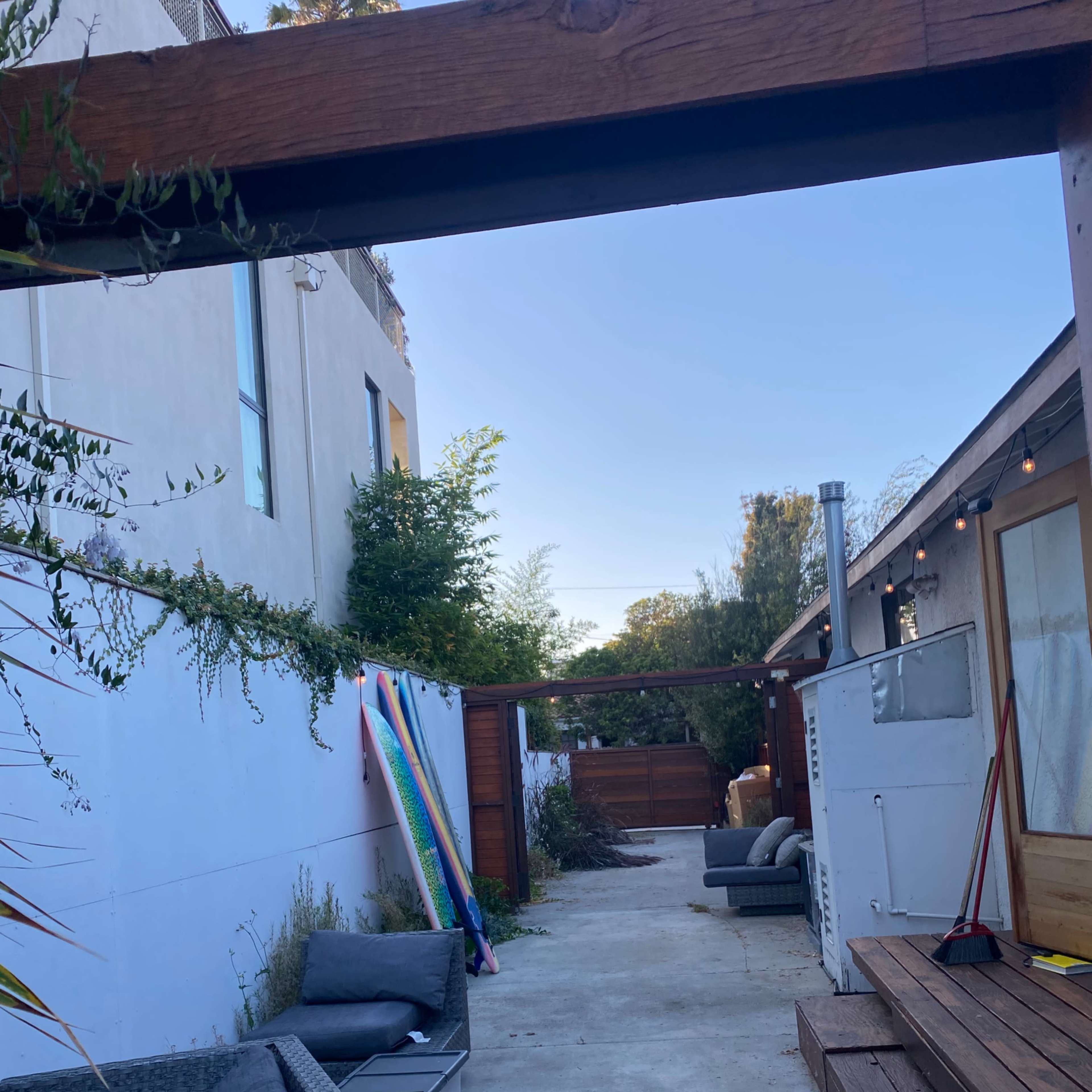 A narrow outdoor pathway lined with potted plants, leading towards a wooden fence, with seating areas and surfboards stacked against a wall.