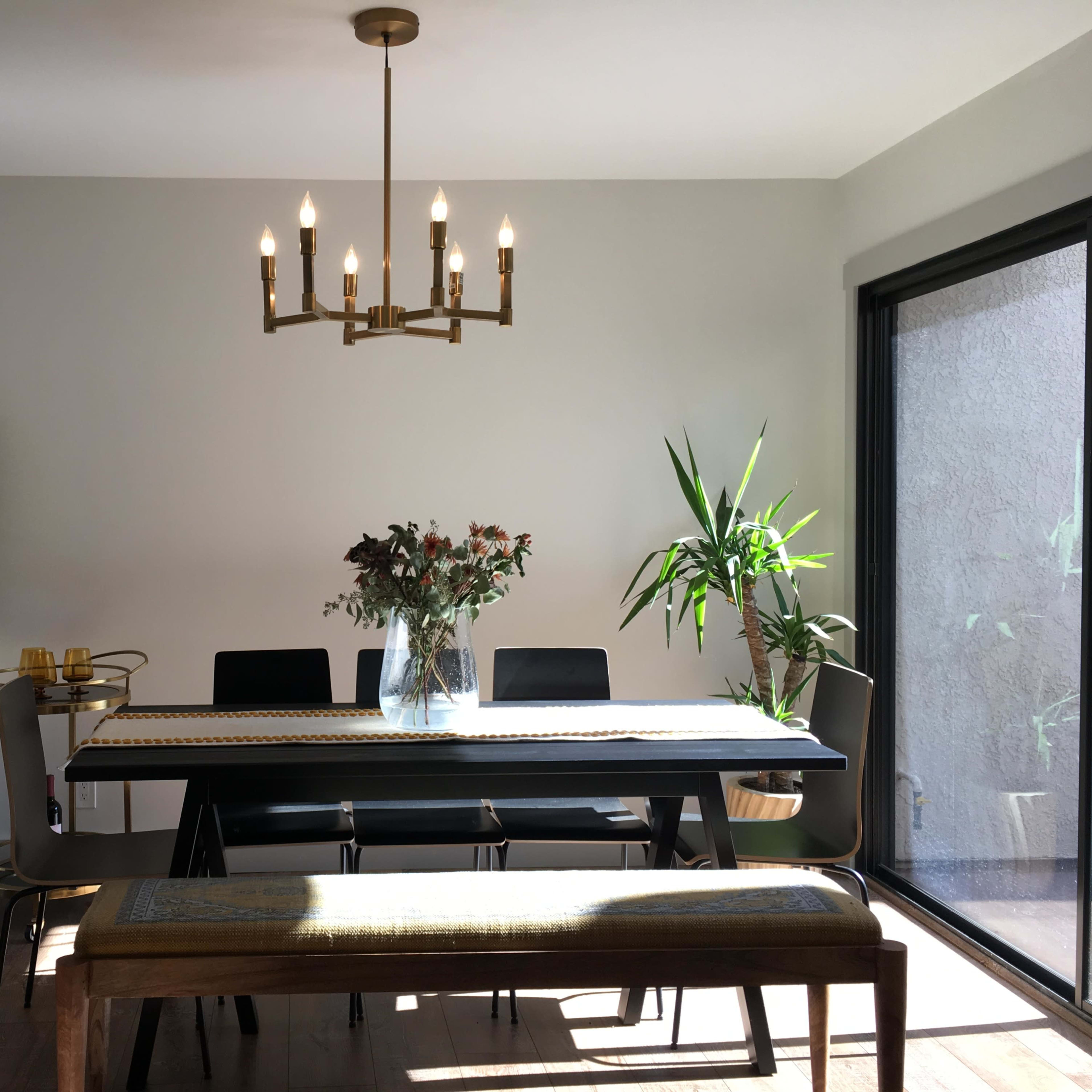 A modern dining area featuring a long table with chairs, a central chandelier, and plants by the window.