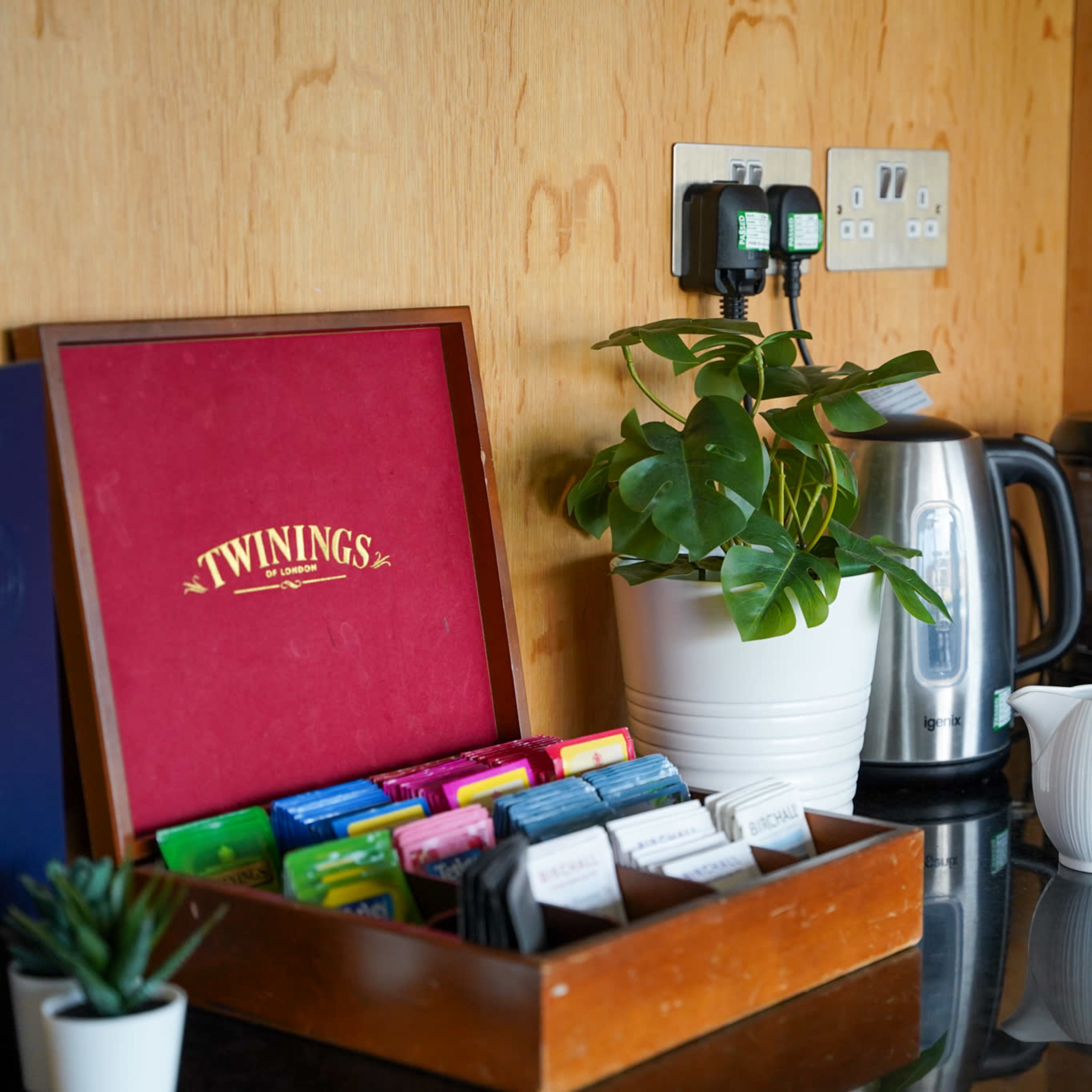 A collection of tea bags is presented in a wooden box labeled "Twinings," next to a kettle, a coffee maker, and two creamers on a black countertop.