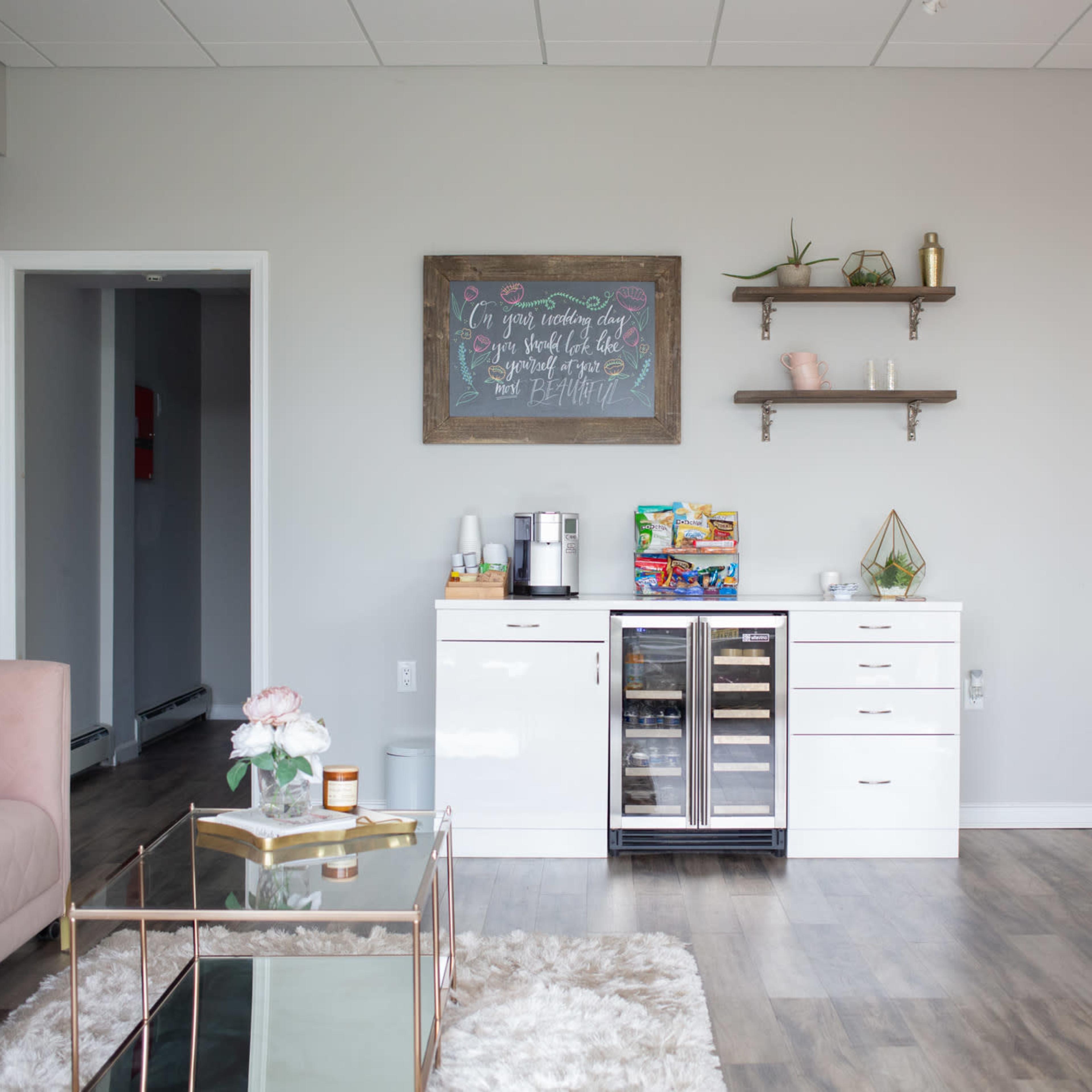 A modern waiting area with a pink sofa, a coffee machine, a beverage refrigerator, and decorative shelves against a light gray wall.