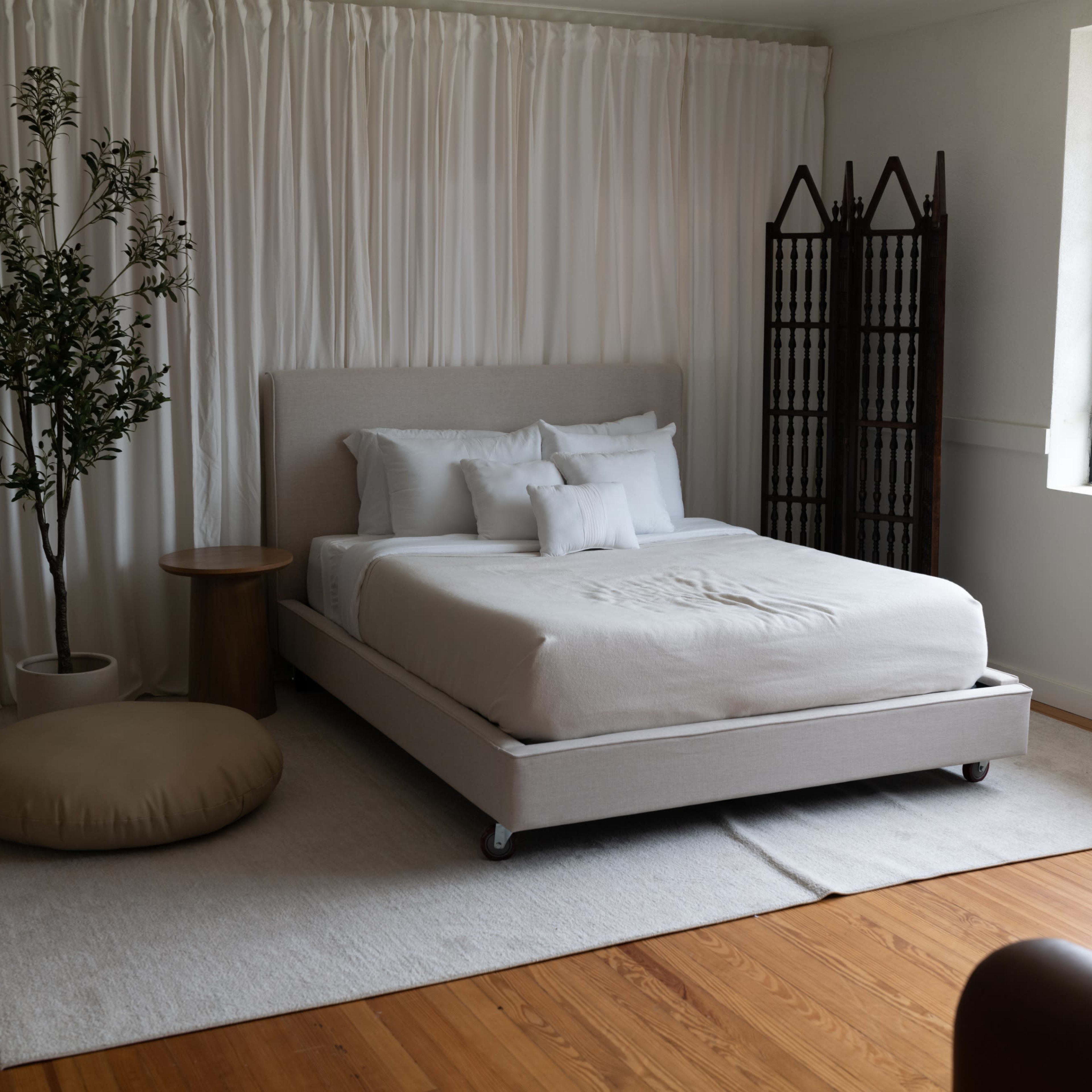 A minimalist bedroom featuring a bed, a potted plant, a round ottoman, and a folding screen against a light wall with large windows.