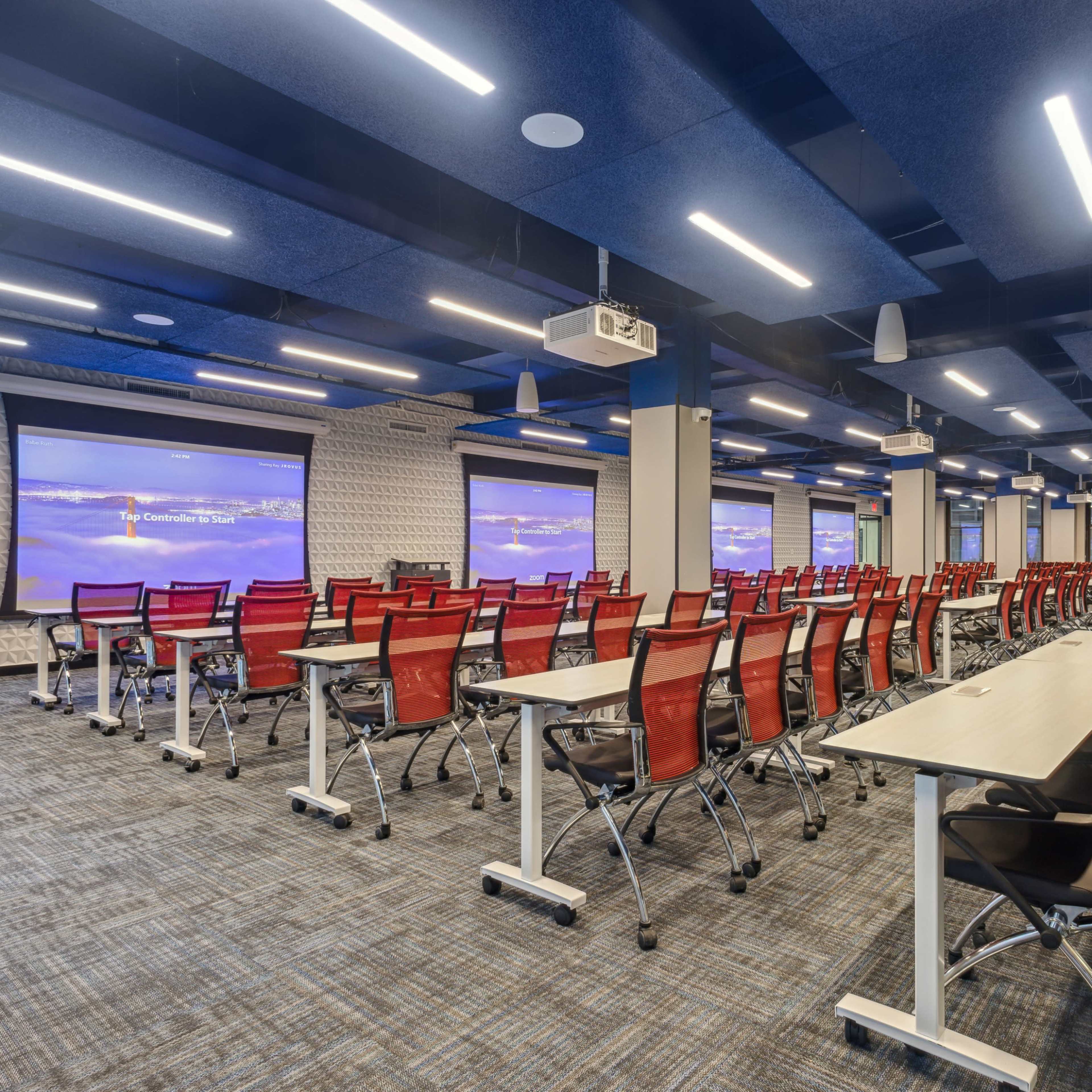 A large conference room features rows of tables and red chairs, with two projection screens at the front.