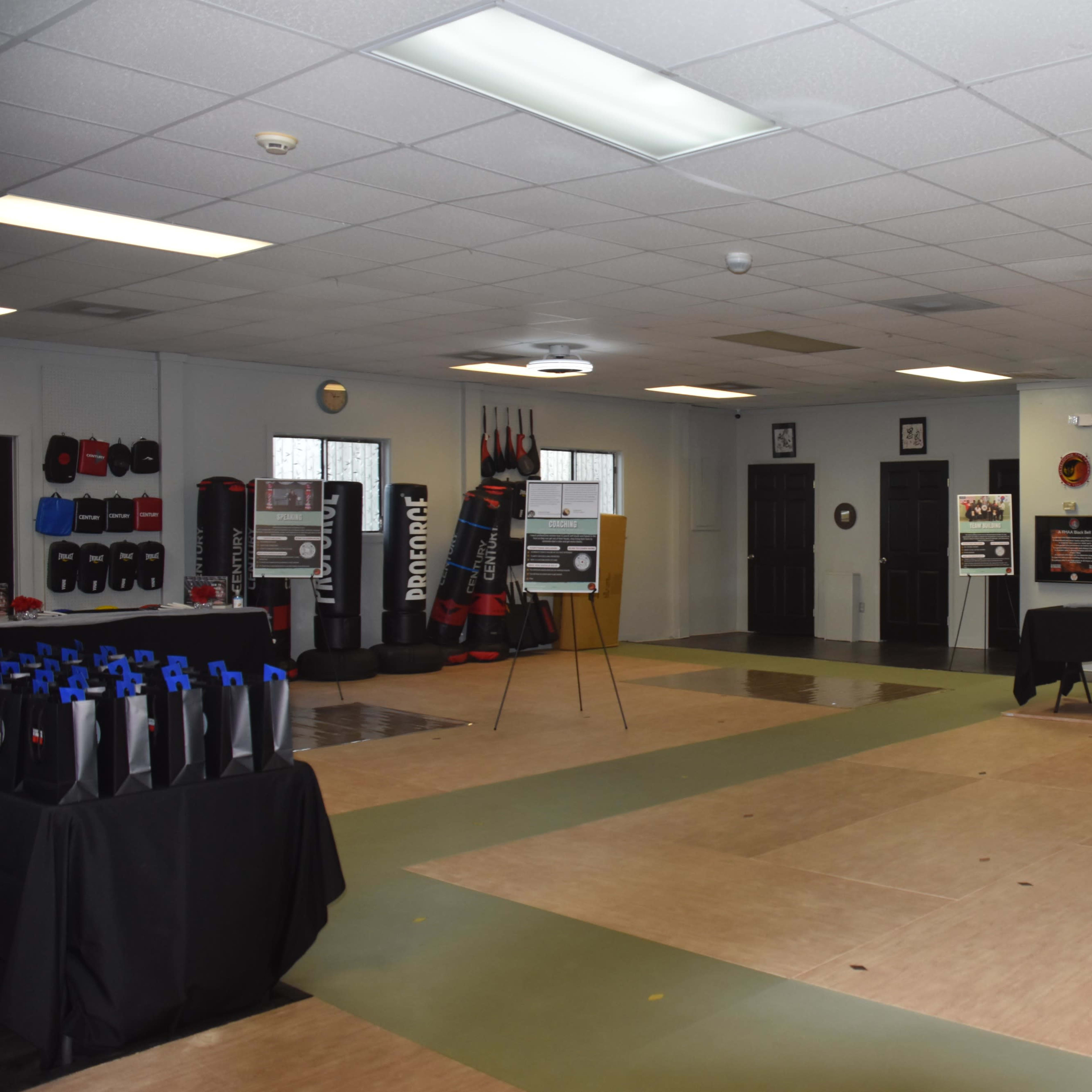 A spacious training room with martial arts equipment, black tables set up for presentations, and padded walls adorned with training gear.