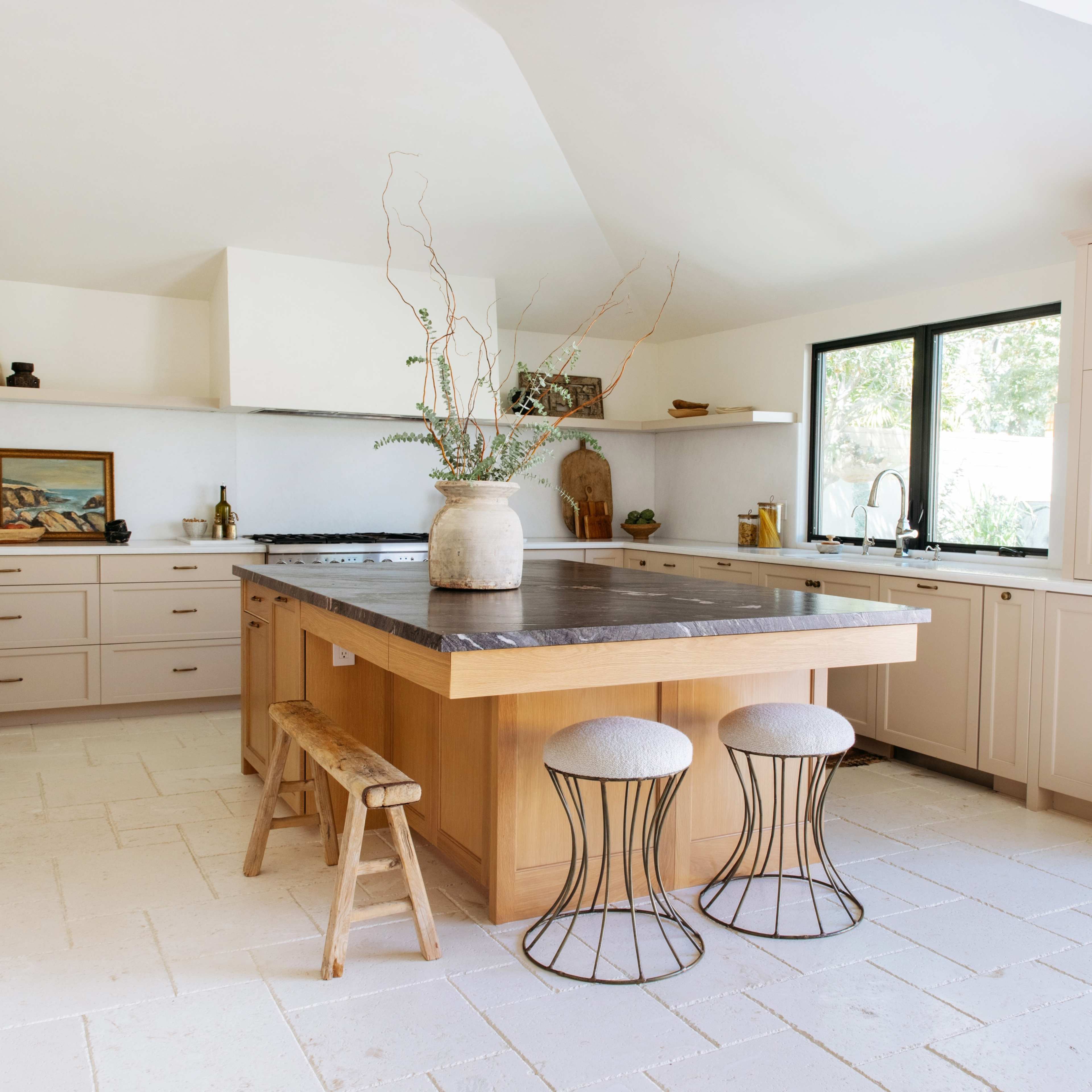 A modern kitchen with a central wooden island, two metal stools, and cabinetry on either side, featuring light-colored walls and a large window.